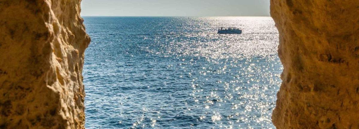 A scenic view of a bright blue ocean with sparkling sunlight on the water, framed by two rough tan stone walls in the foreground, with a small boat visible in the distance.