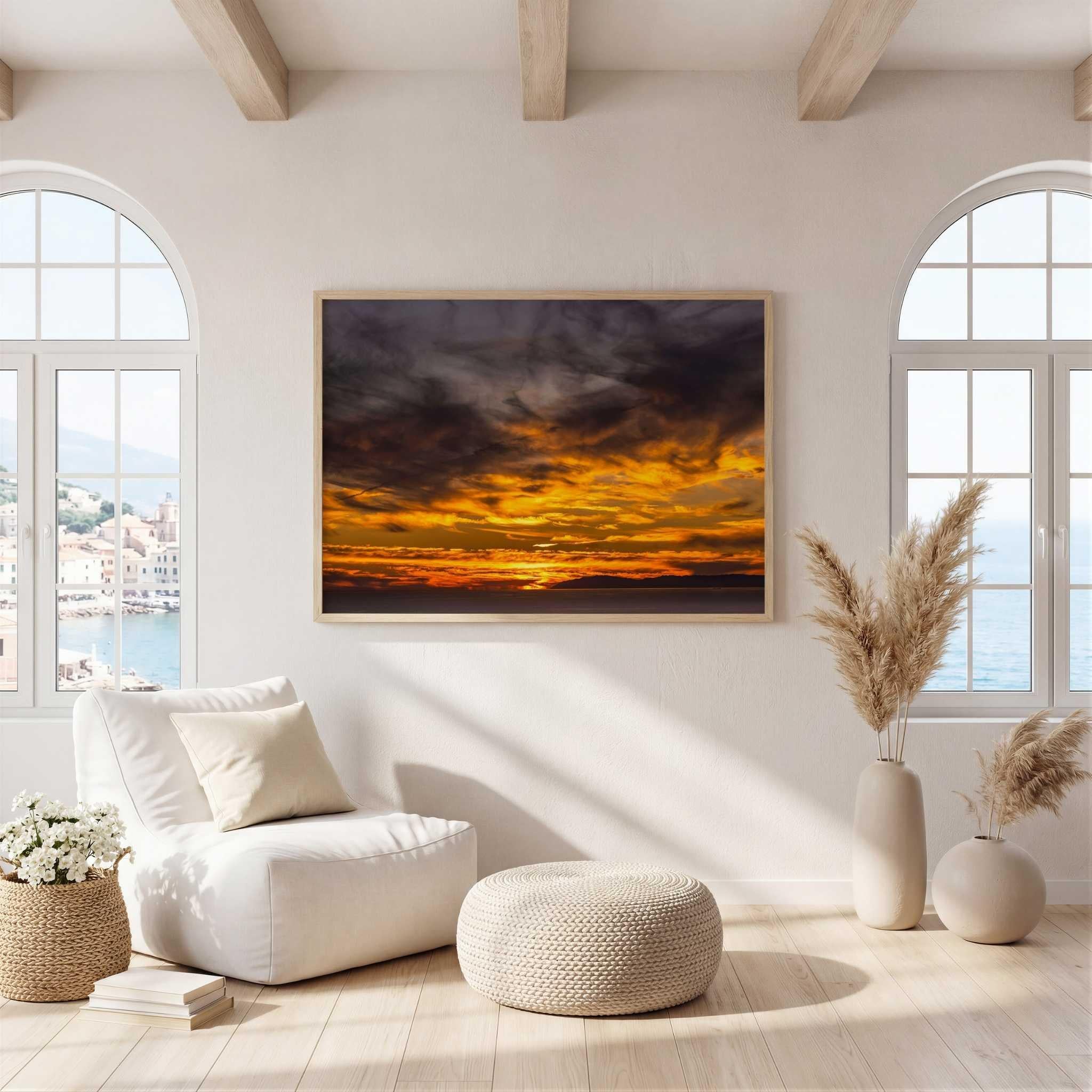 A framed wall art print of gleaming golden sunset, in Laguna Beach ,showing a dramatic orange and yellow sunset over dark clouds, displayed in a bright, modern living room with a white armchair, ottoman, and arched windows.