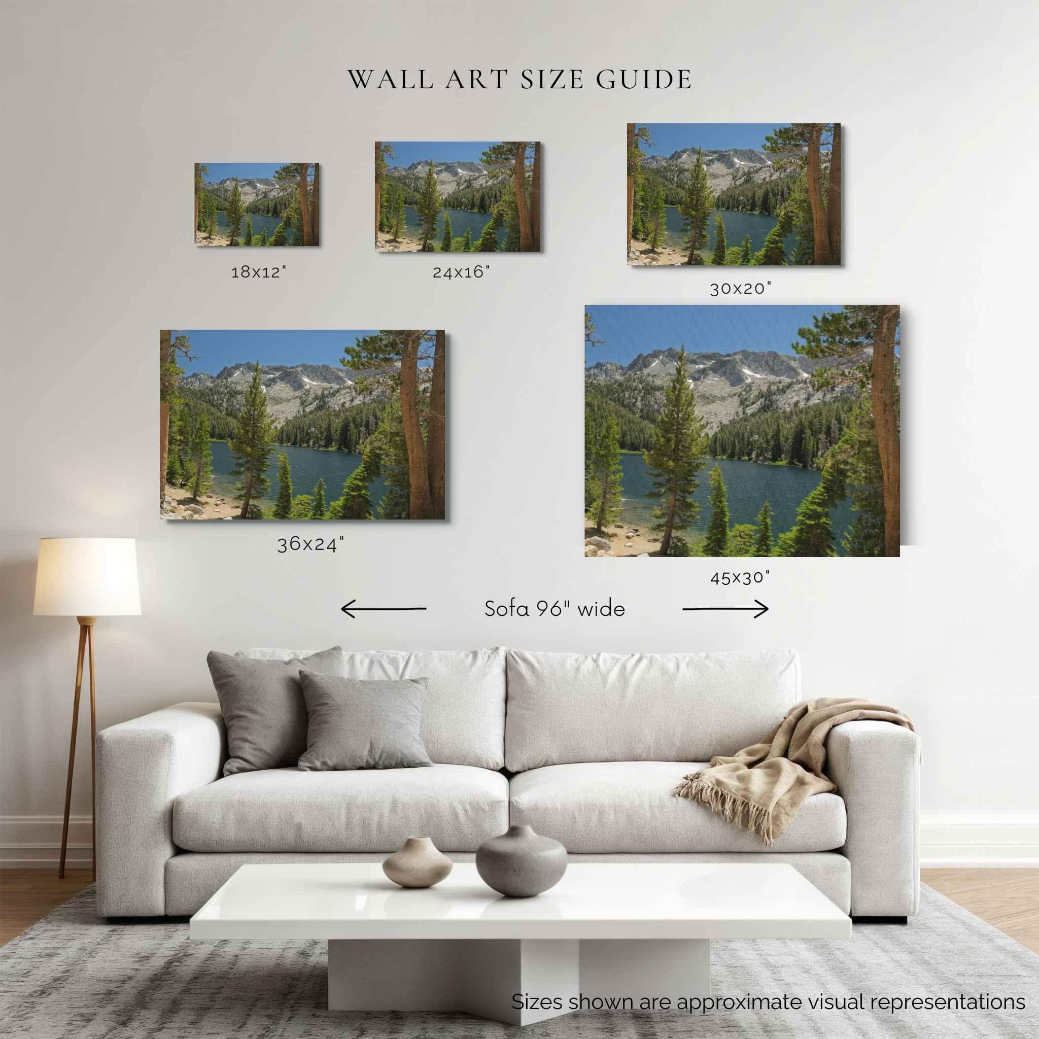 A staged living room showing a wall-mounted set of six landscape wall art prints of where the trail meets the mountains, with size labels and arrows comparing print dimensions to a sofa width. The room includes a light gray sofa, white coffee table, floor lamp, and rug.