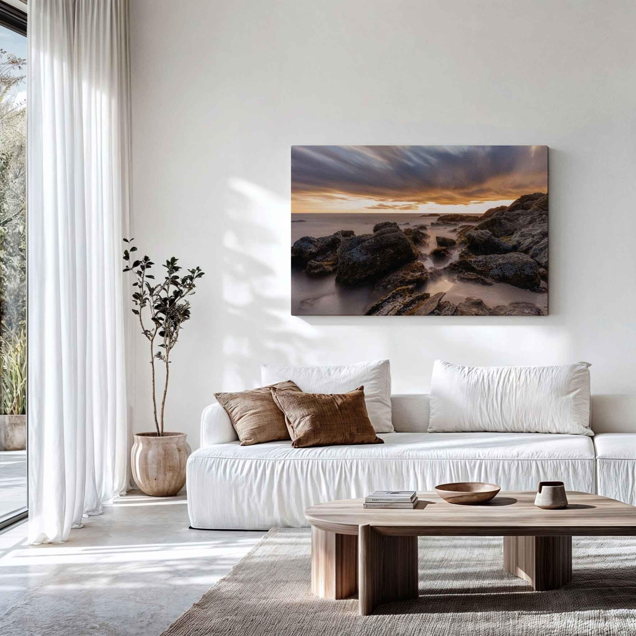 After the Storm canvas print displayed above a white sofa in a modern living room, featuring a dramatic long exposure sunset over the rocky shoreline of Laguna Beach, California.