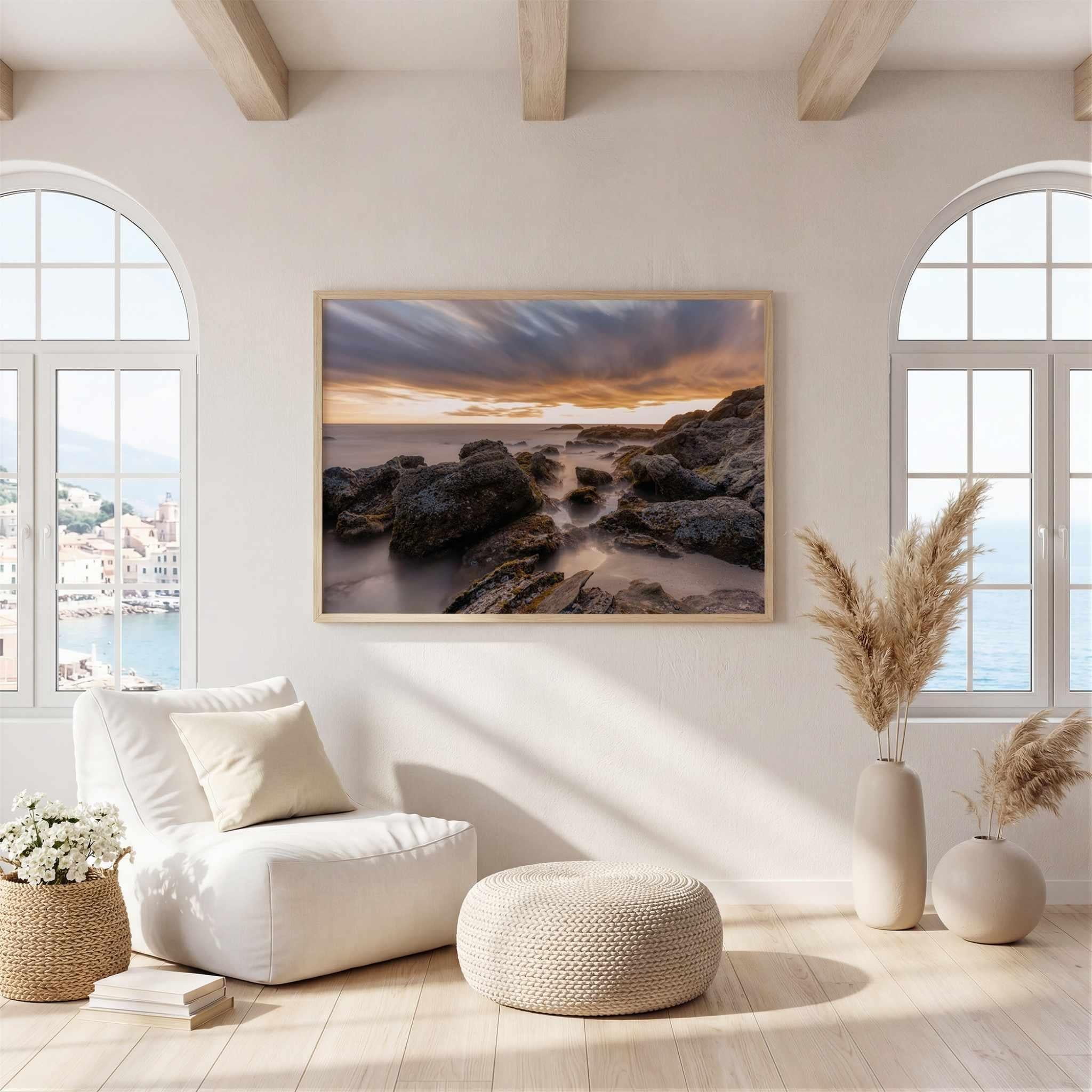 Laguna Beach Coastal framed landscape art print depicting a rocky coastline at sunset, displayed on a light beige wall in a bright, modern living room with a white armchair, ottoman, and pampas grass vases.