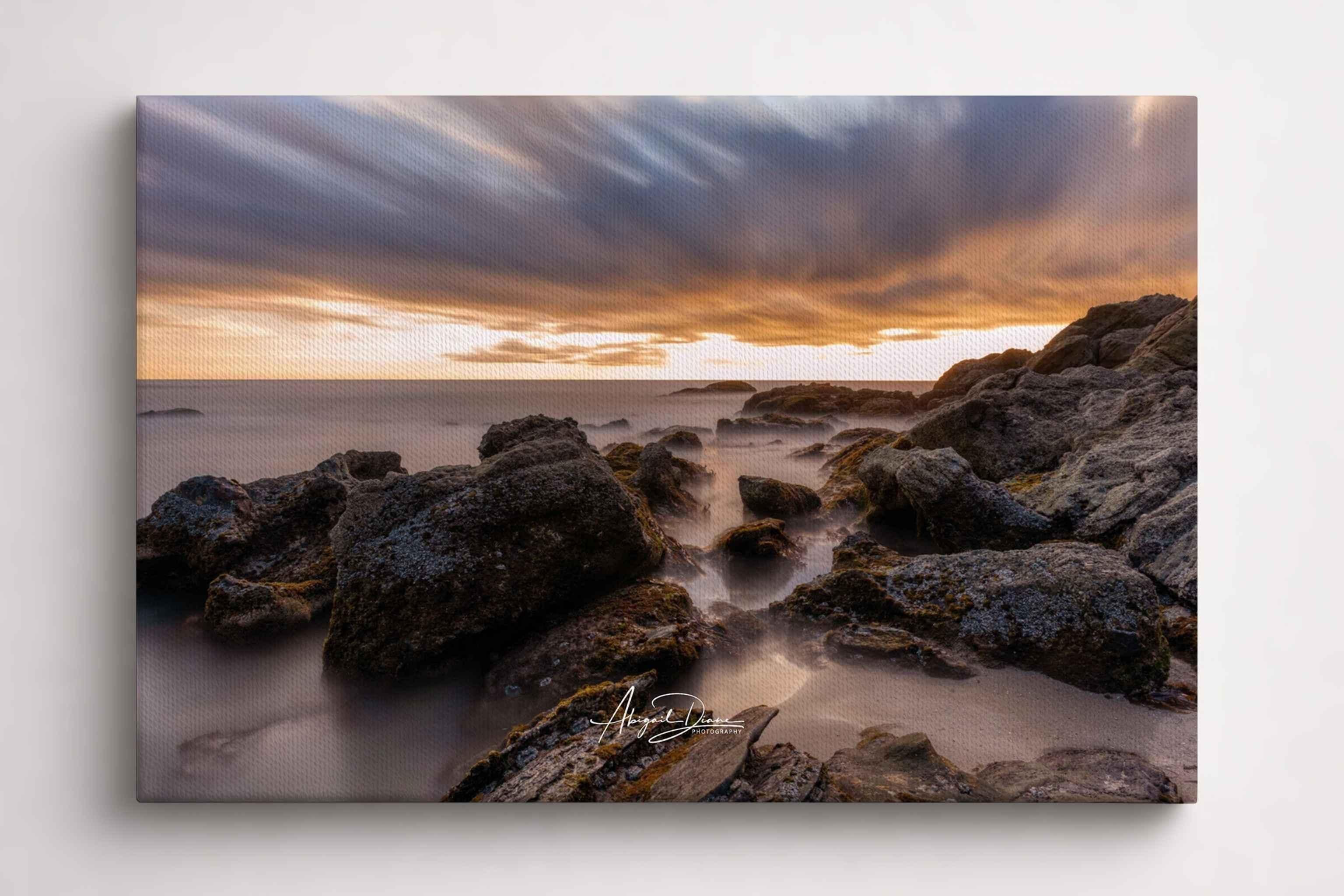 Gallery wrapped canvas print of After the Storm featuring a dramatic coastal sunset over rugged shoreline rocks, displayed in a front side-angle mockup highlighting the premium canvas texture, gallery wrap, and rich warm tones by Abigail Diane Photography.