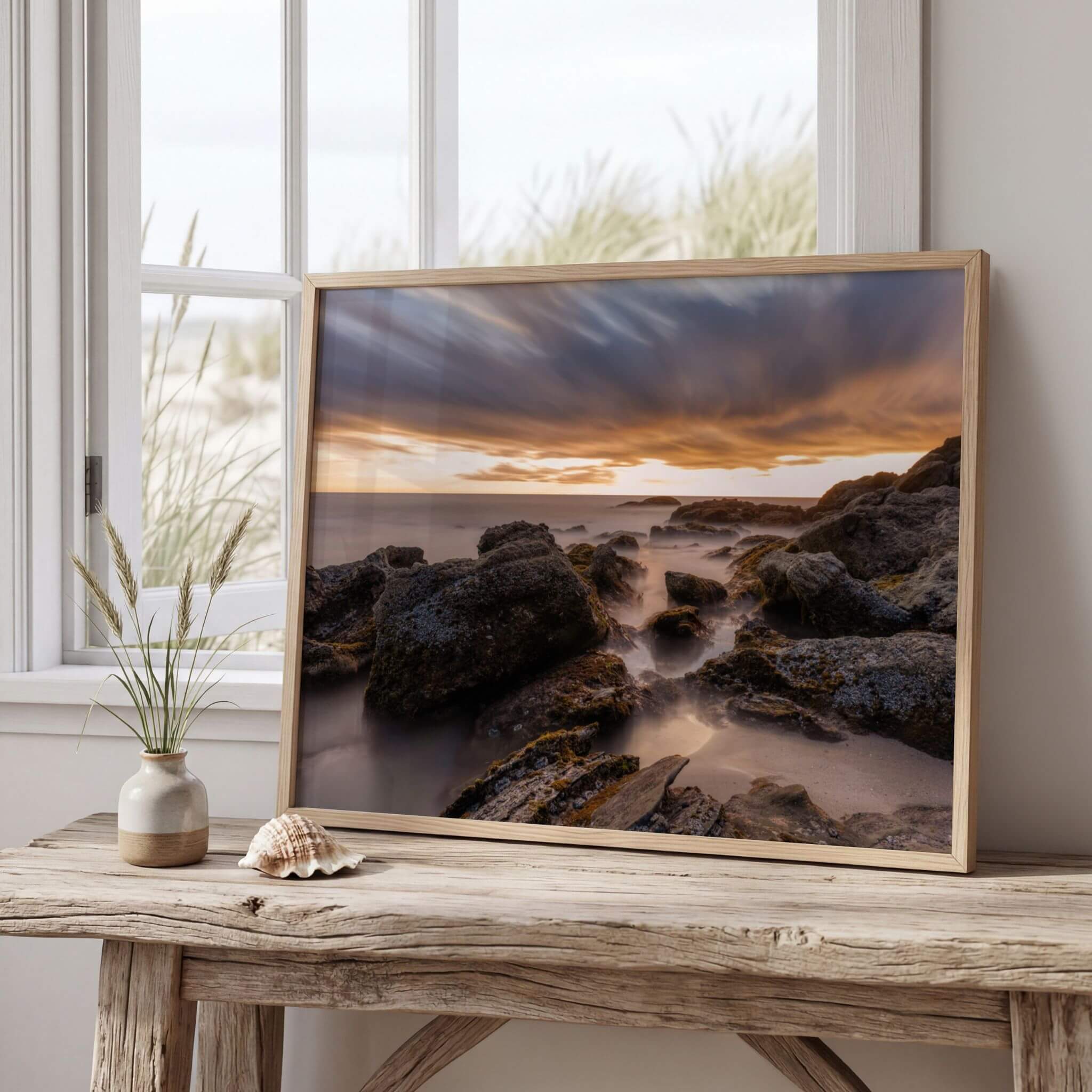 Framed Laguna Beach coastal landscape print displayed on a wooden console beside a seaside window, featuring rugged shoreline rocks, long exposure waves, and glowing sunset clouds.