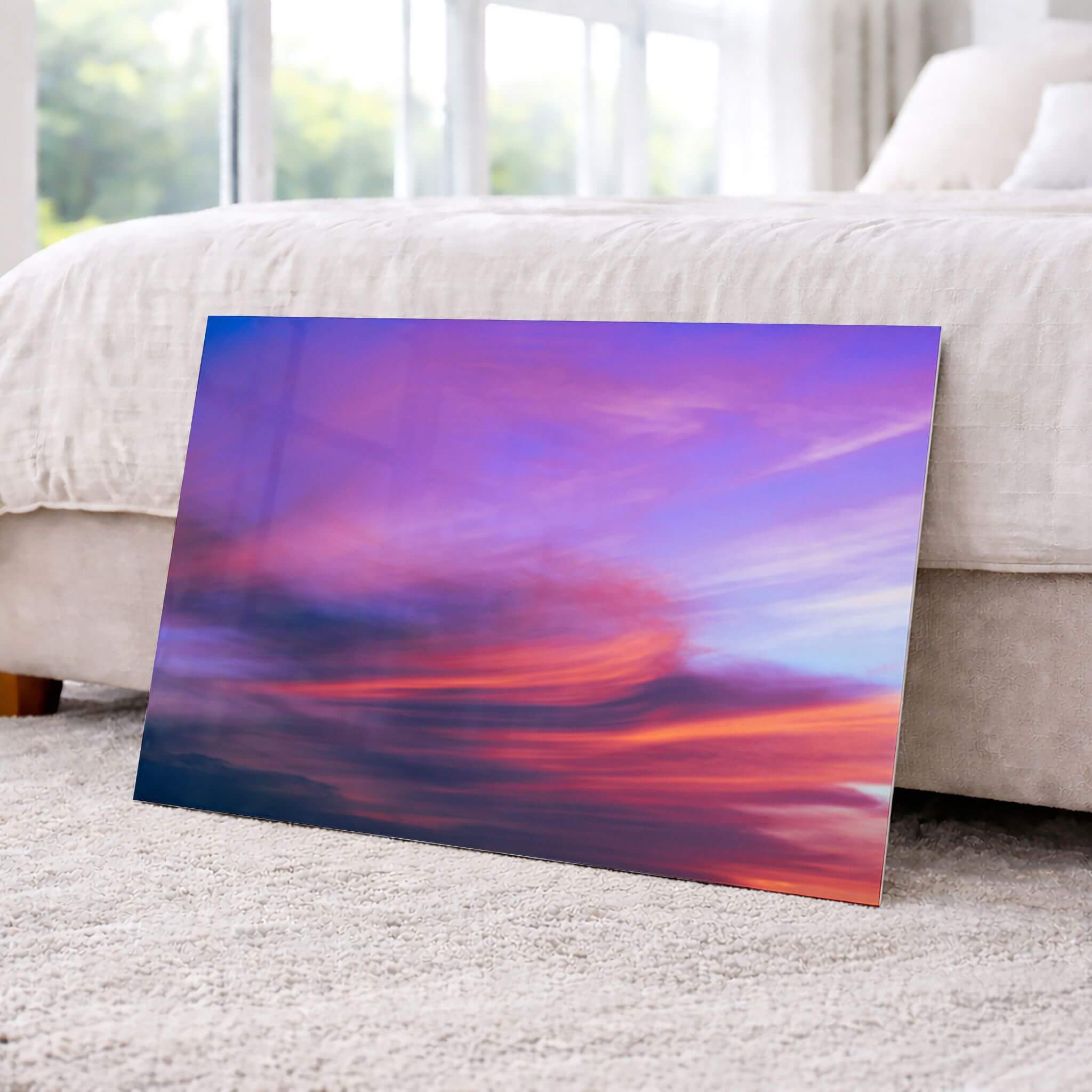 Lavender Dreams HD metal wall art leaning against a bed in a bright bedroom, featuring flowing lavender, coral, pink, and blue sunset clouds by Abigail Diane Photography.