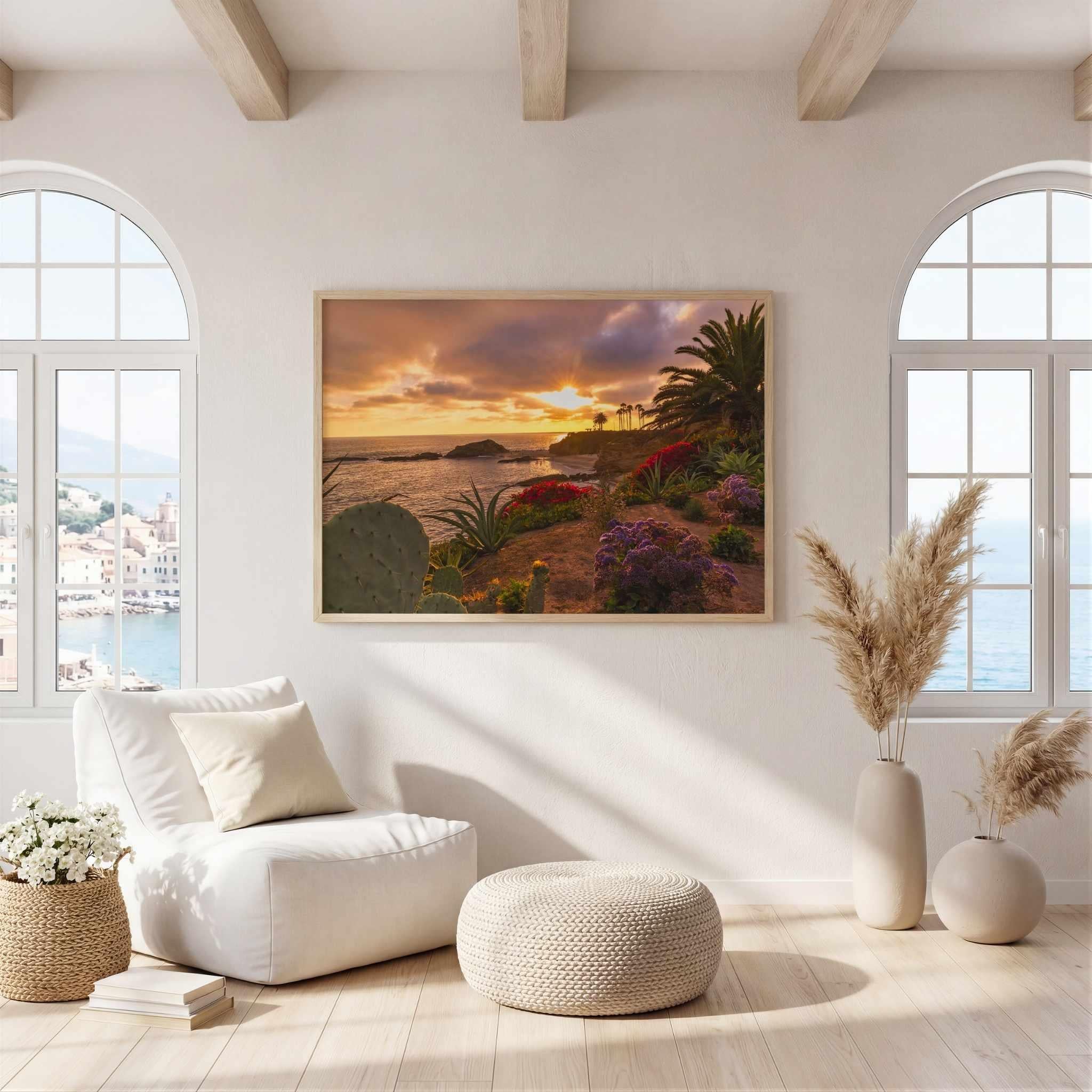 A framed landscape art print depicting a tropical sunset beach scene with palm trees and purple flowers, displayed on a white wall in a modern, sunlit living room with a white armchair, ottoman, and decorative vases.