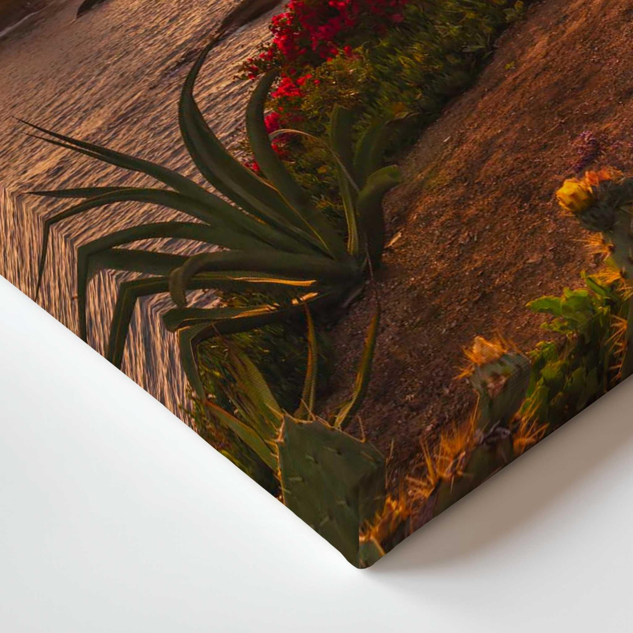 A rectangular wall art print or canvas shown at an angle against a white wall. The printed scene depicts desert plants, including a large green agave-like plant, cacti, and red flowers on a brown textured background.