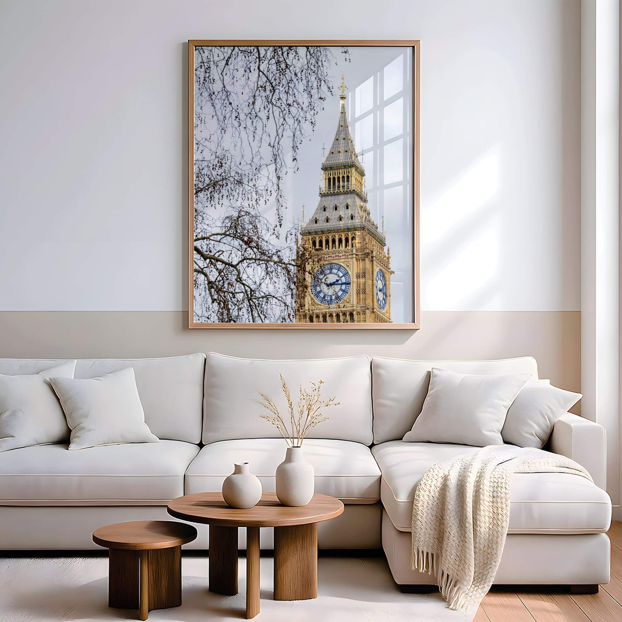 A large framed wall art print of Big Ben hanging above a modern white sectional sofa in a bright, neutral living room. The print features a light wood frame and shows the clock tower against a pale sky with bare tree branches.