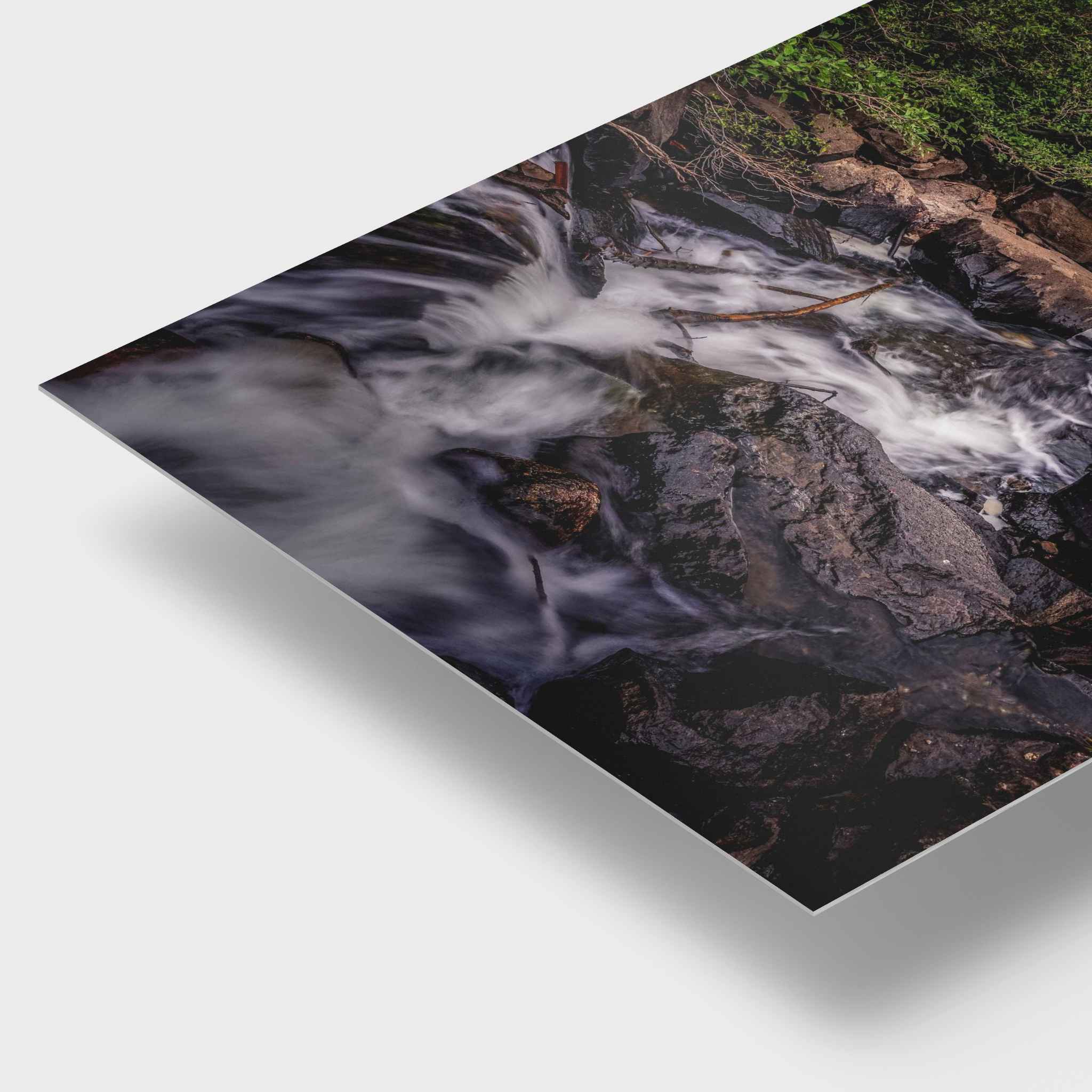 Cascading into stillness HD Metal corner print showing a close-up photograph of a rushing stream over dark rocks, displayed at an angle on a light gray background with a drop shadow.
