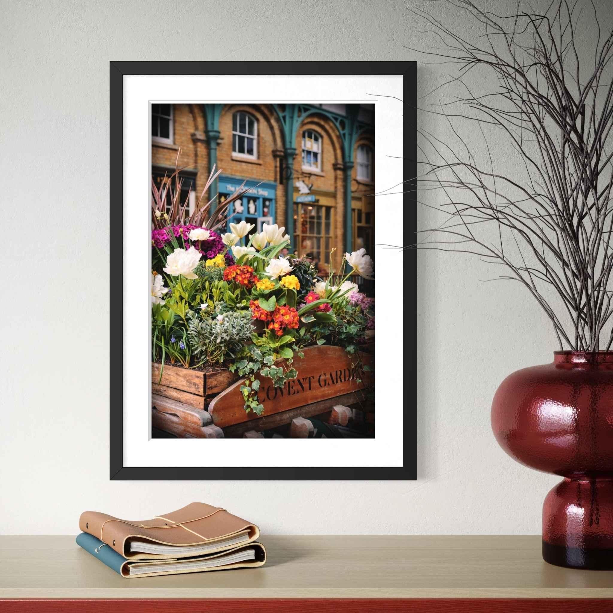 Black framed fine art print with white mat showcasing Covent Garden in Bloom by Abigail Diane Photography. Vibrant spring flowers and historic London architecture are displayed on a styled tabletop with books and a decorative red vase.