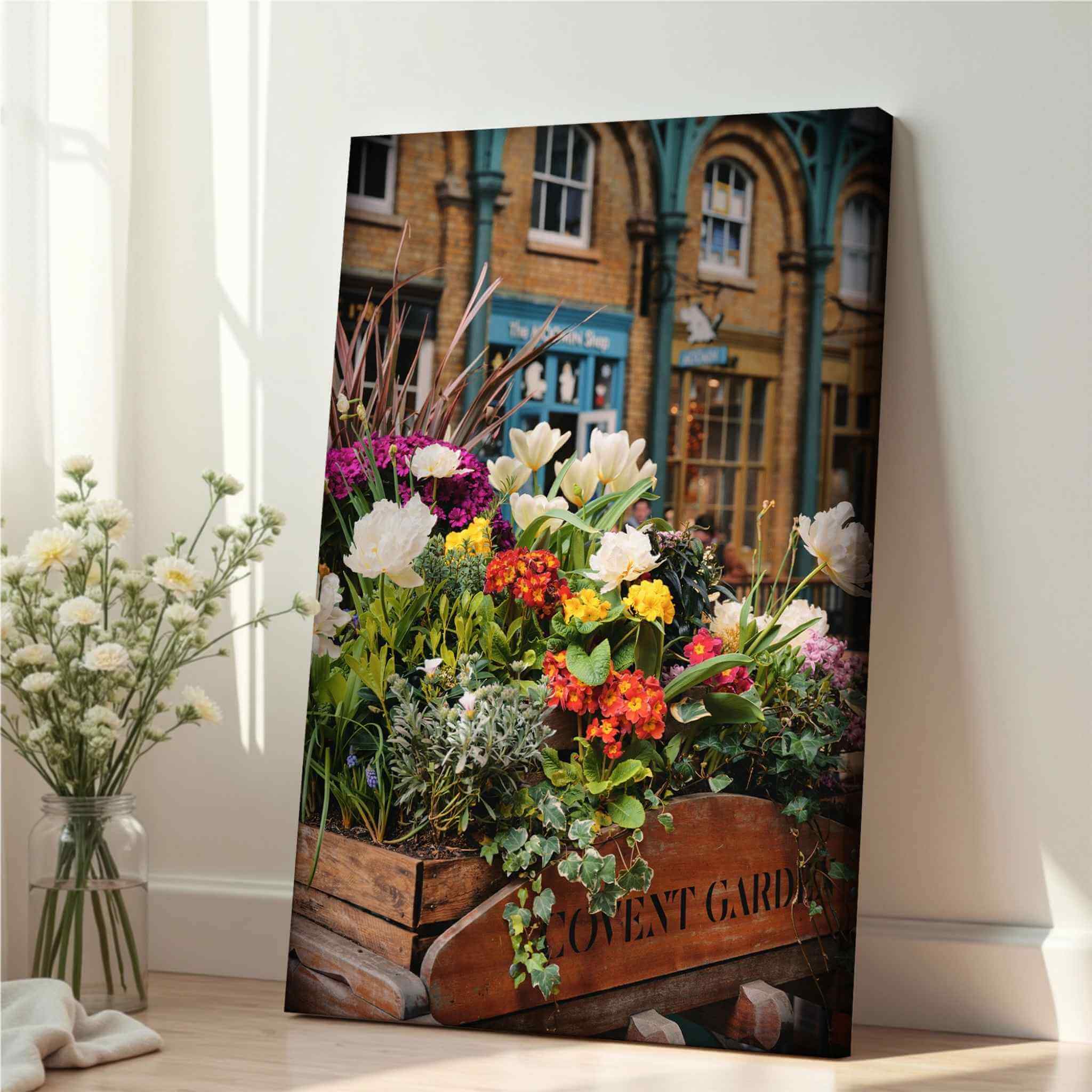 Covent Garden in Bloom by Abigail Diane Photography displayed as a vertical canvas print leaning against a bright interior wall, featuring colourful seasonal flowers and the historic Covent Garden Market in London, England