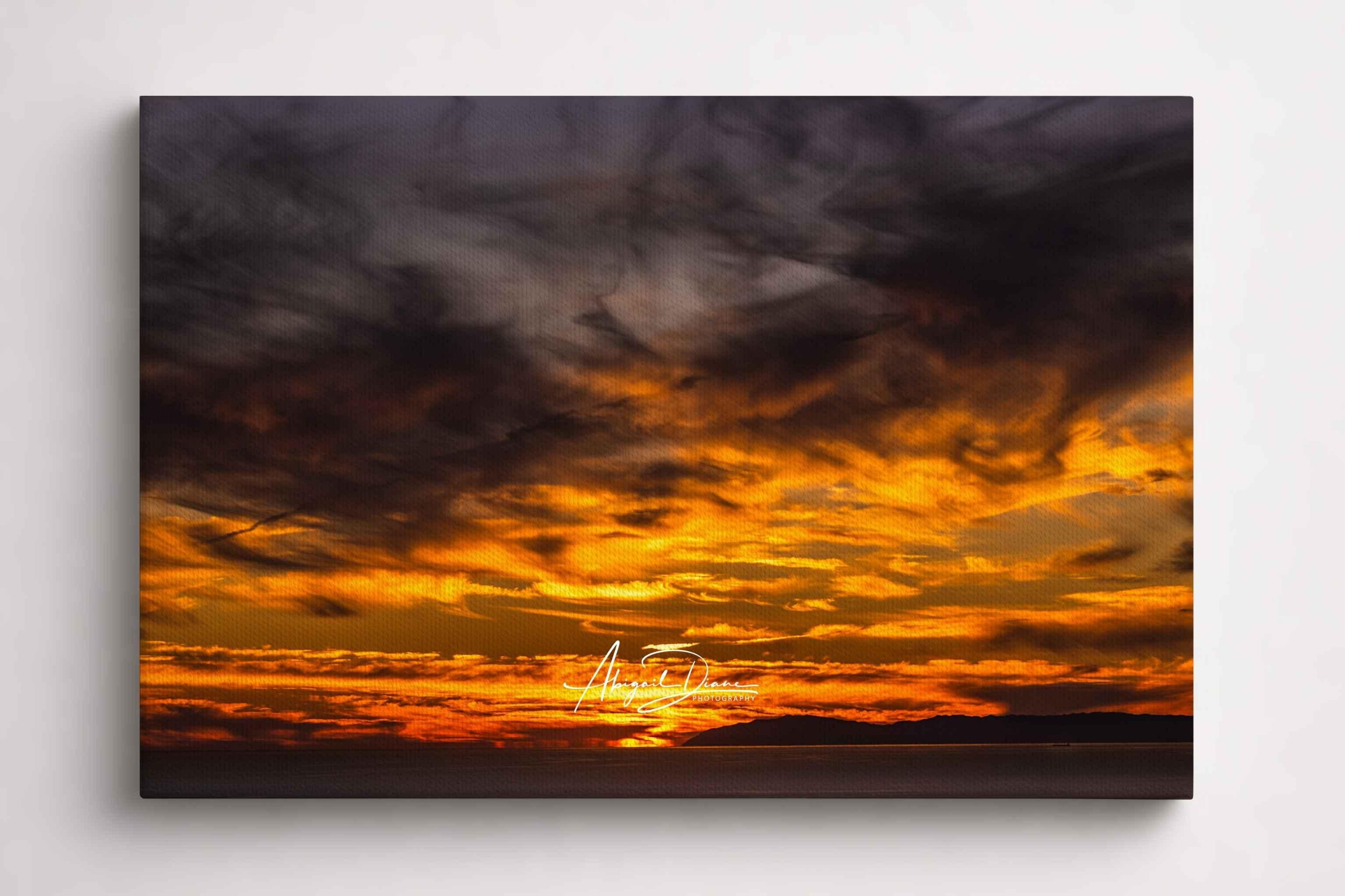 Golden sunset over Catalina Island in the Pacific Ocean showing a dramatic sunset sky with dark gray clouds, bright orange and yellow light near the horizon, and a dark silhouette along the bottom. Gallery-wrapped canvas coastal print by Abigail Diane Photography.