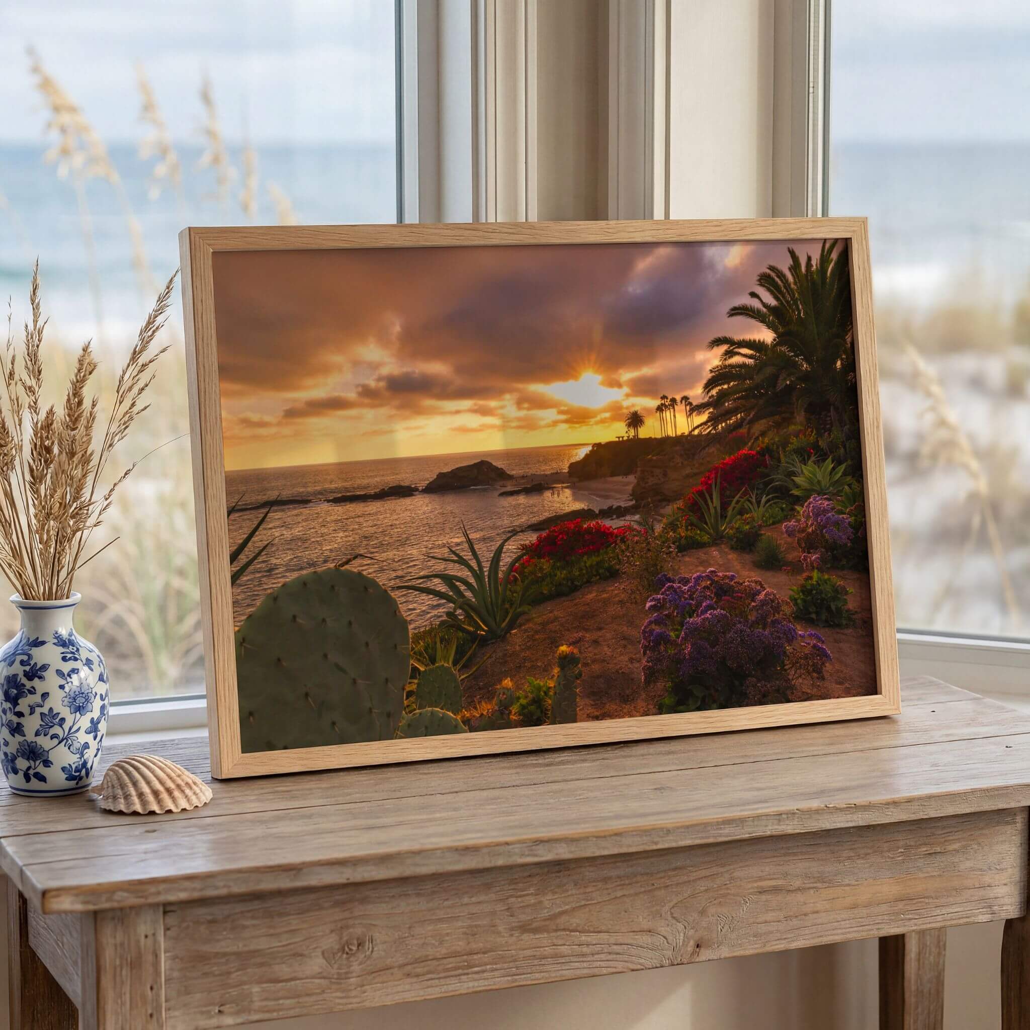 Laguna Beach sunset wall art displayed on a wooden console table beside a beach window, featuring vibrant coastal flowers, palm trees, and a glowing Pacific Ocean sunset..