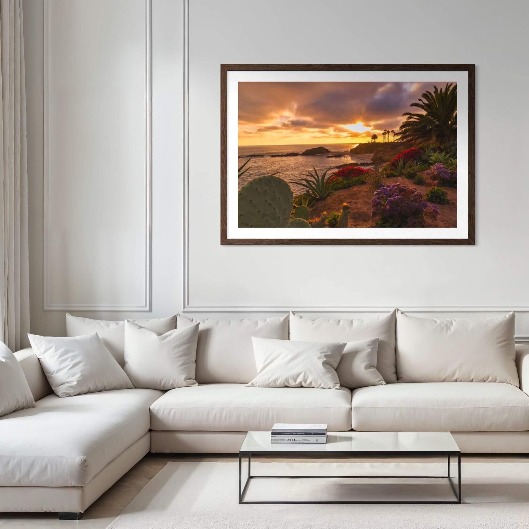 Framed Laguna Beach sunset fine art print displayed above a contemporary sofa, capturing golden evening light, coastal flowers, palm trees, and the Pacific Ocean.