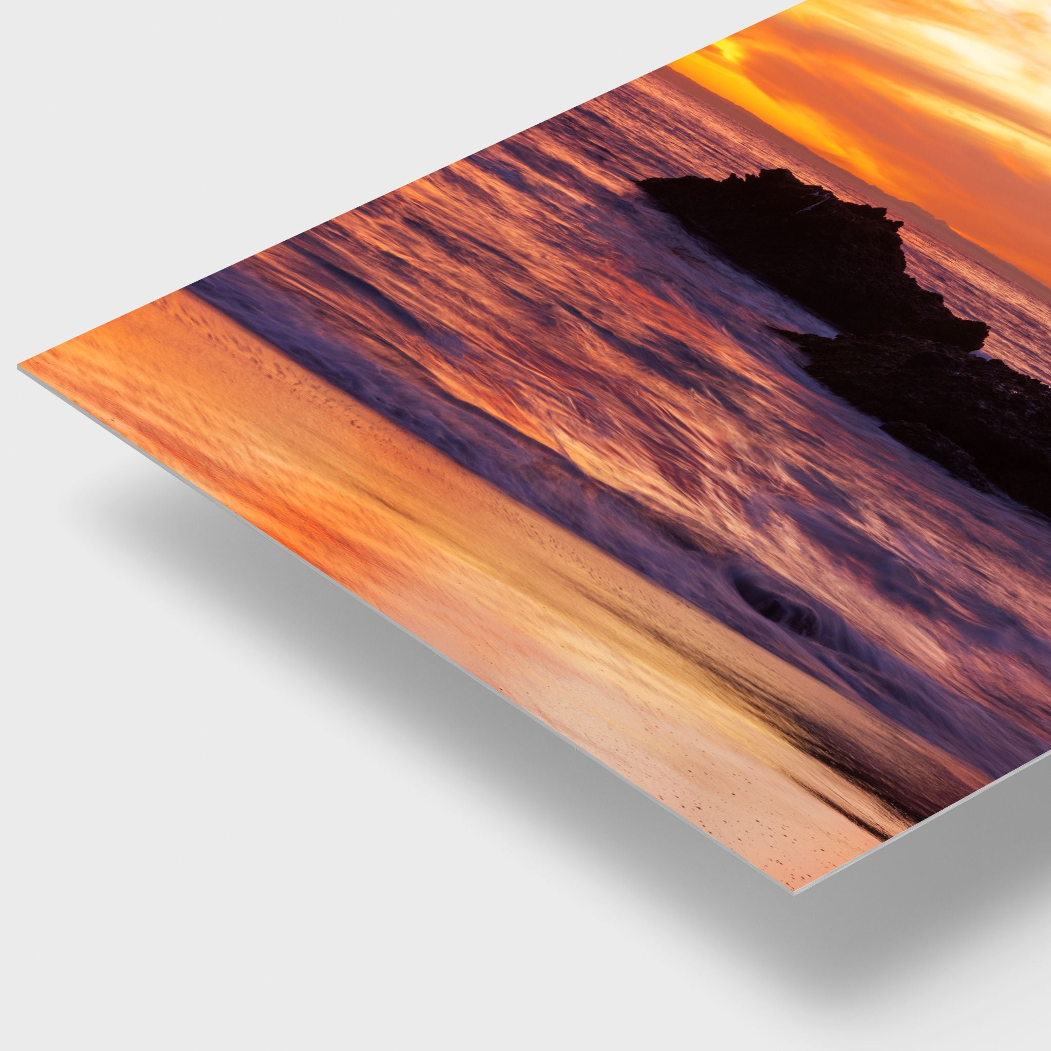 A close up metal art print showing a dramatic sunset over water with orange and purple tones, displayed at an angle on a soft light background with a drop shadow.
