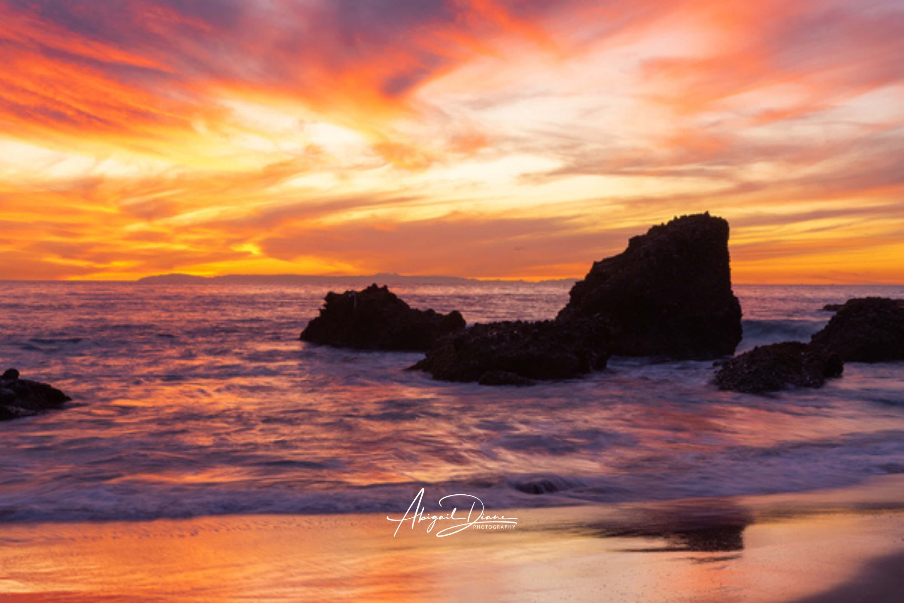Vibrant sunset over Woods Cove Beach in Laguna Beach California featuring glowing sherbet-toned skies in shades of orange, pink, gold, and purple above ocean waves and dark coastal rock formations, photographed by Abigail Diane Photography.