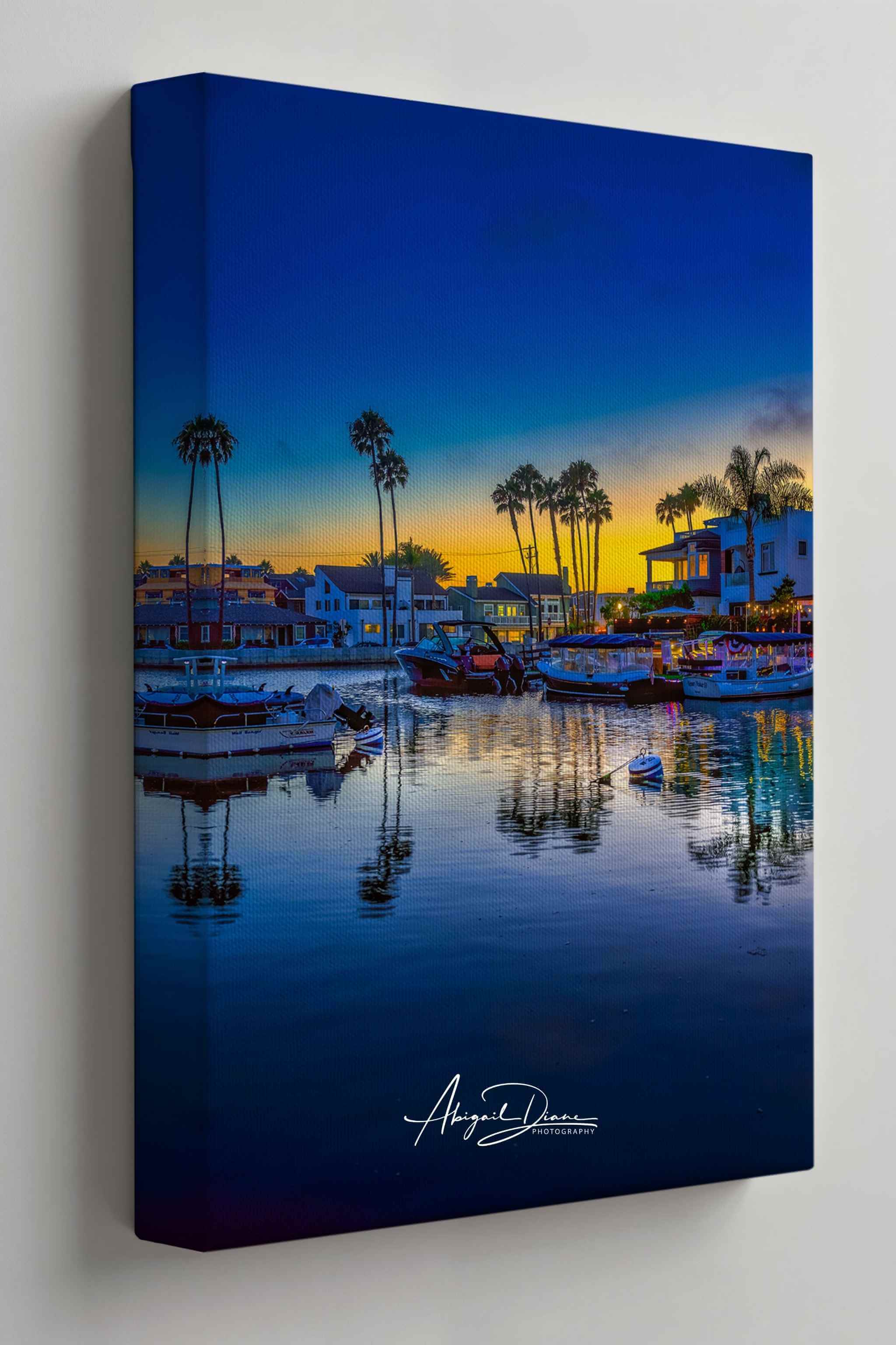 A vertical canvas wall art print depicting a Newport Beach harbor scene at sunset, with boats on calm water, palm trees, buildings, and a deep blue sky. The canvas is shown at a slight angle against a light gray wall. Photography by Abigail Diane Photography