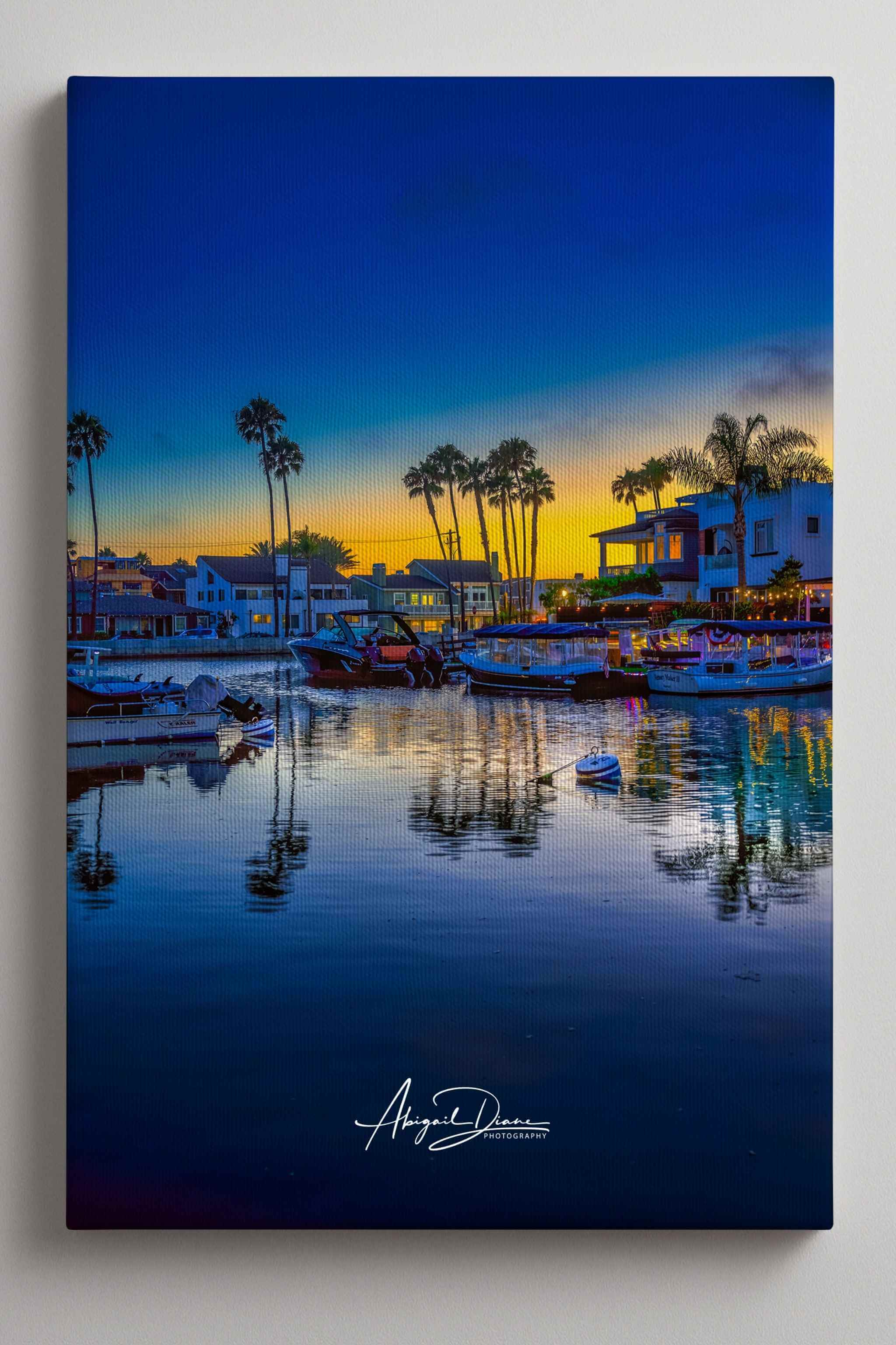 A vertical canvas wall art print depicting a Newport Beach harbor scene at sunset, with boats on calm water, palm trees, buildings, and a deep blue sky. The canvas is shown at a slight angle against a light gray wall. Photography by Abigail Diane Photography
