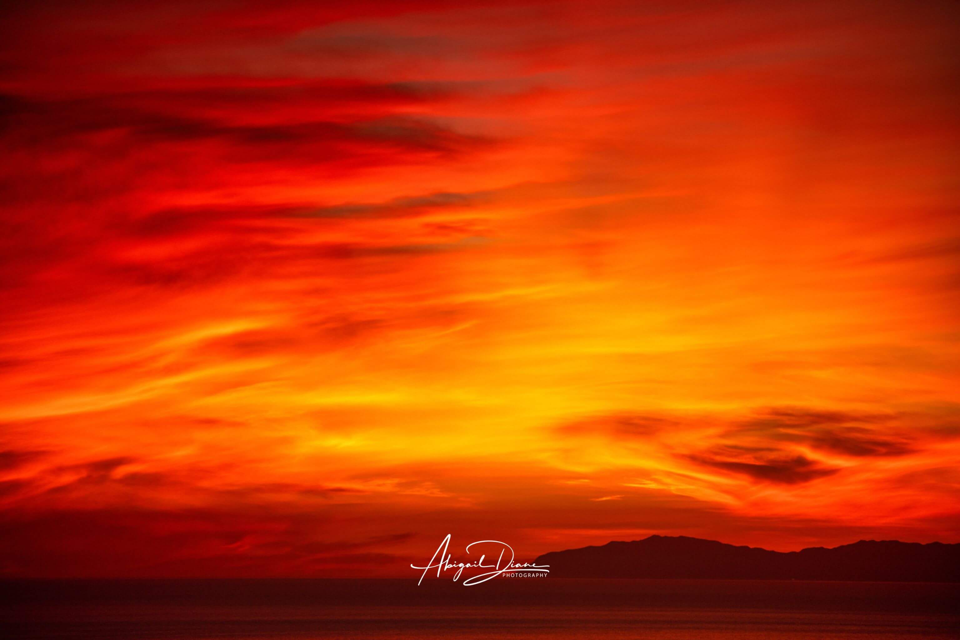 Inferno Sunset over Catalina Island with fiery red, orange and golden clouds glowing above the Pacific Ocean off the California coast. Photographed by Abigail Diane Photography.