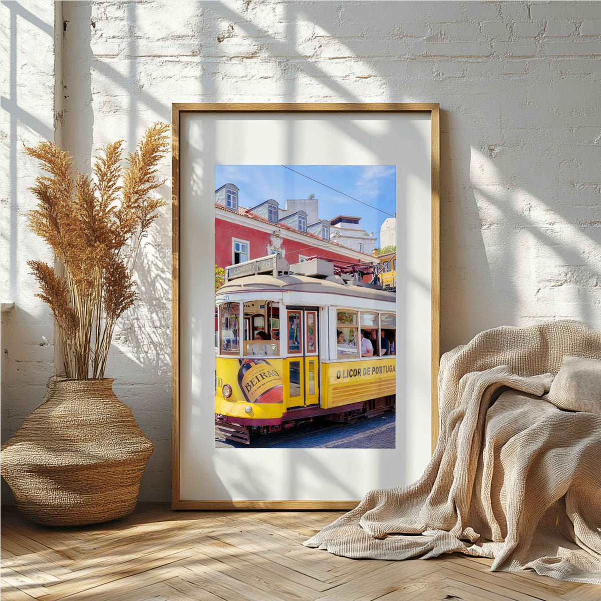 A framed art print of a yellow Lisbon tram on a city street, leaning against a white brick wall in a styled room with a woven vase of dried grass and a beige throw blanket.