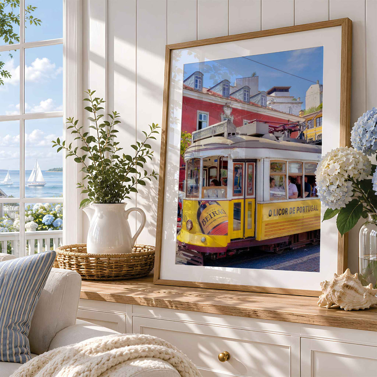 A framed art print featuring a vintage yellow Lisbon tram on a colorful street, styled in a bright, modern room next to a window with a potted plant and flowers.