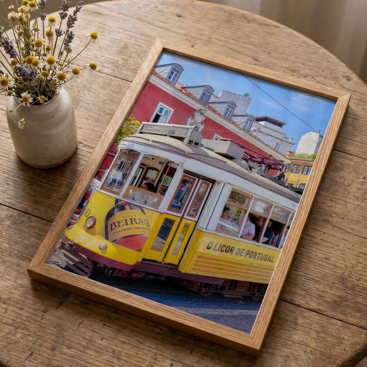 A rectangular framed art print featuring a vintage yellow tram on a wooden street, styled on a rustic wooden table next to a small ceramic vase with dried yellow flowers.