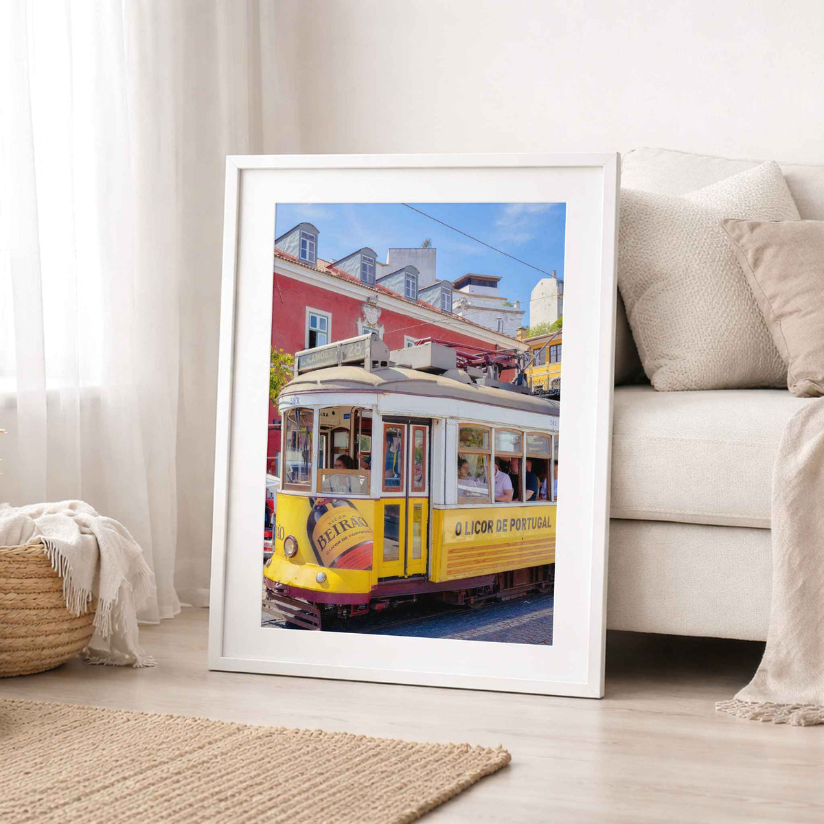 A framed art print leaning against a white sofa in a bright, modern living room. The print features a colorful photograph of a yellow vintage tram on a street with red buildings under a blue sky.
