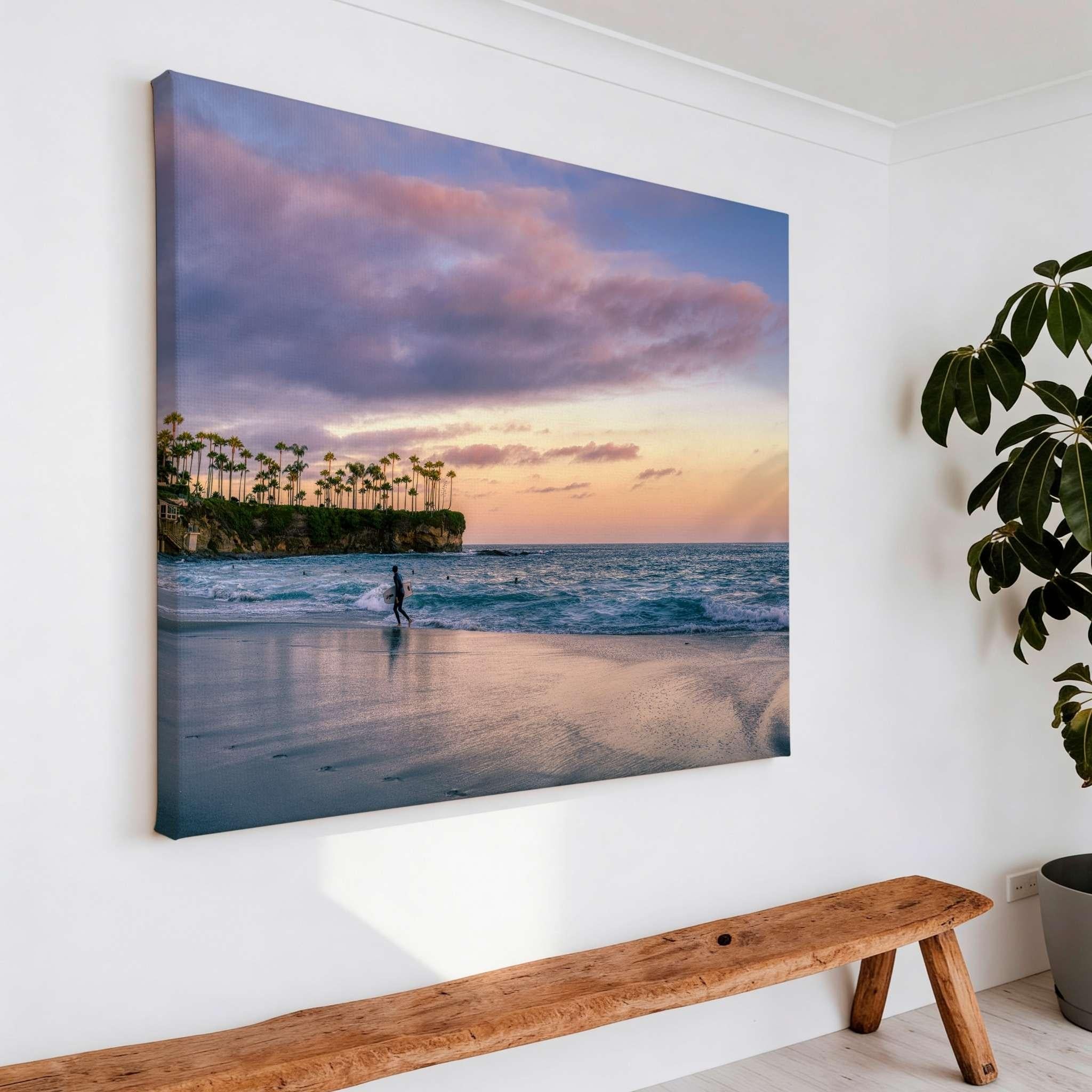 Large gallery-wrapped canvas print of Surfing a California Sunset by Abigail Diane Photography displayed in a modern interior above a wooden bench, featuring a surfer walking along Crescent Beach in Laguna Beach, California at sunset.