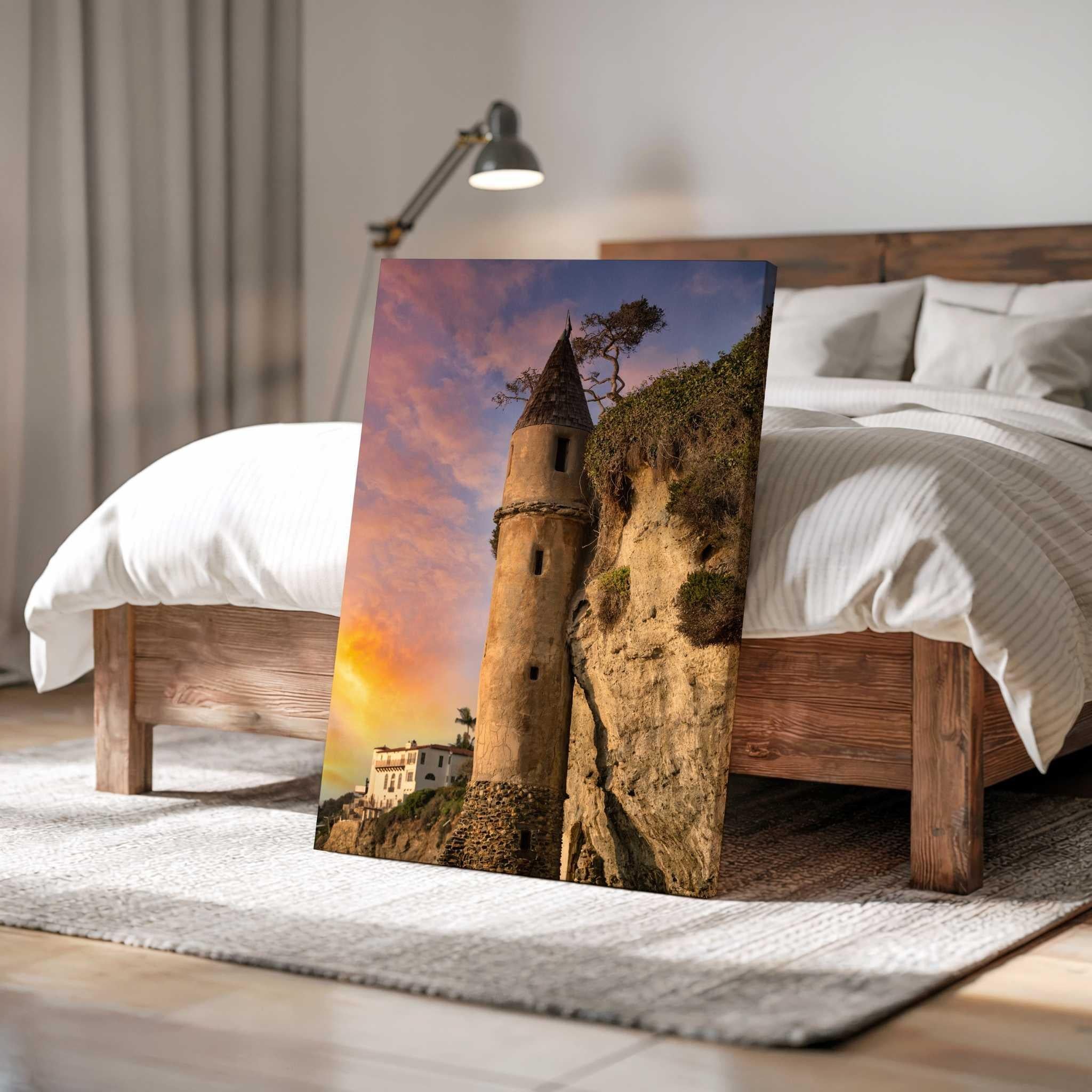 Large gallery wrapped canvas print of Victoria Beach Pirate Tower at sunset in Laguna Beach, California, leaning against a rustic wood bed in a warm neutral bedroom with soft linens and natural light.