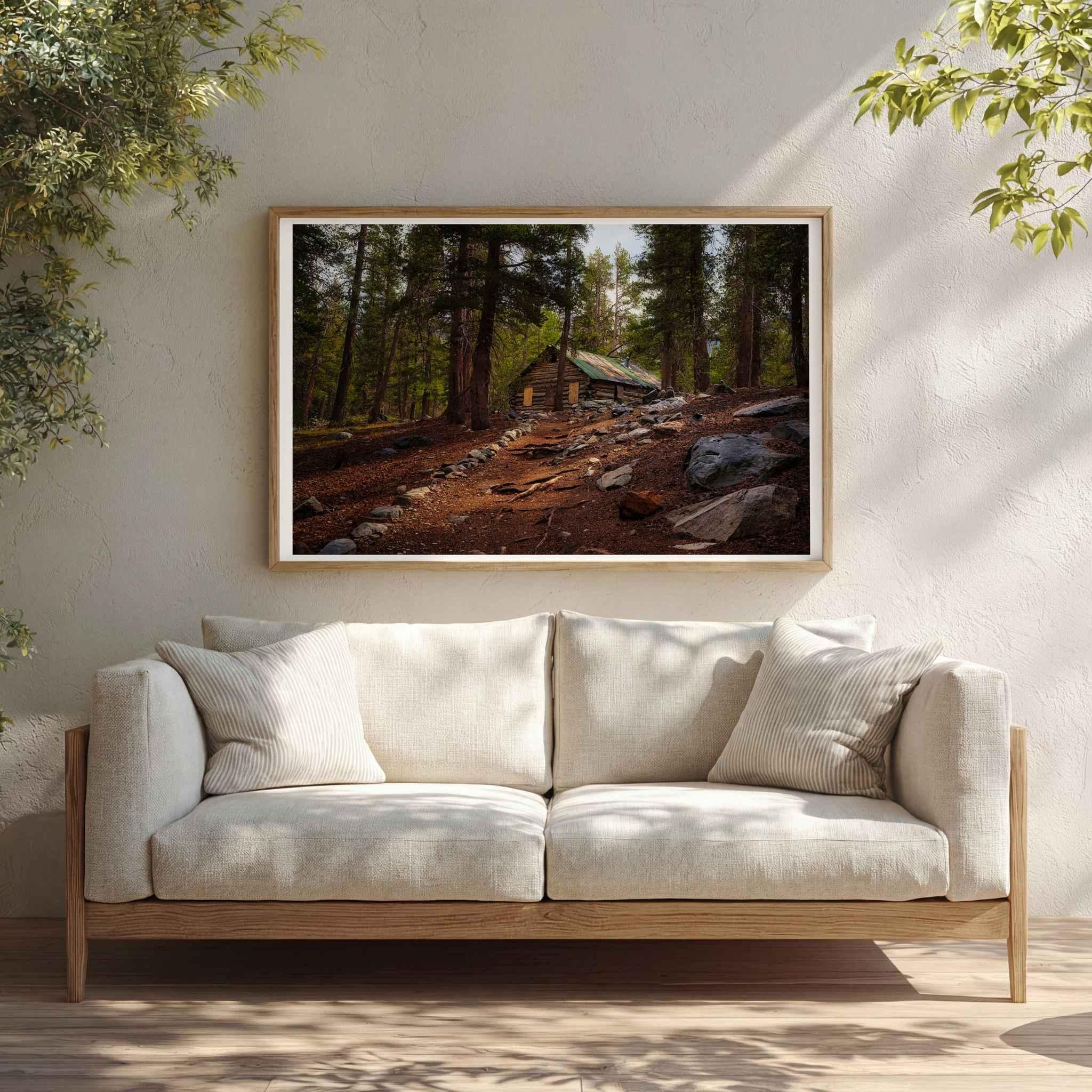 Where the Trail Meets the Past fine art print featuring historic Mahan Cabin near Mammoth Lakes in California’s Eastern Sierra, shown matted in a natural wood frame above a neutral sofa in a sunlit living room.