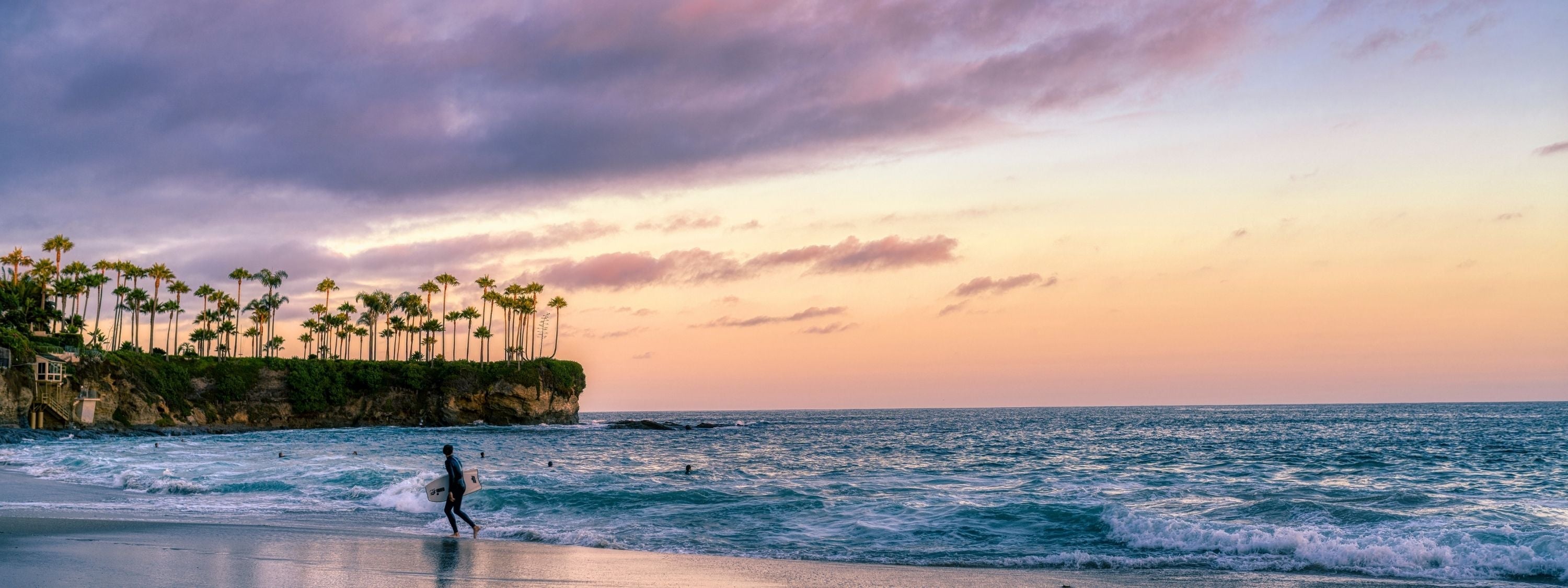Surfing a California Sunset fine art print featuring a lone surfer walking Crescent Beach in Laguna Beach beneath a soft pink Pacific sunset.