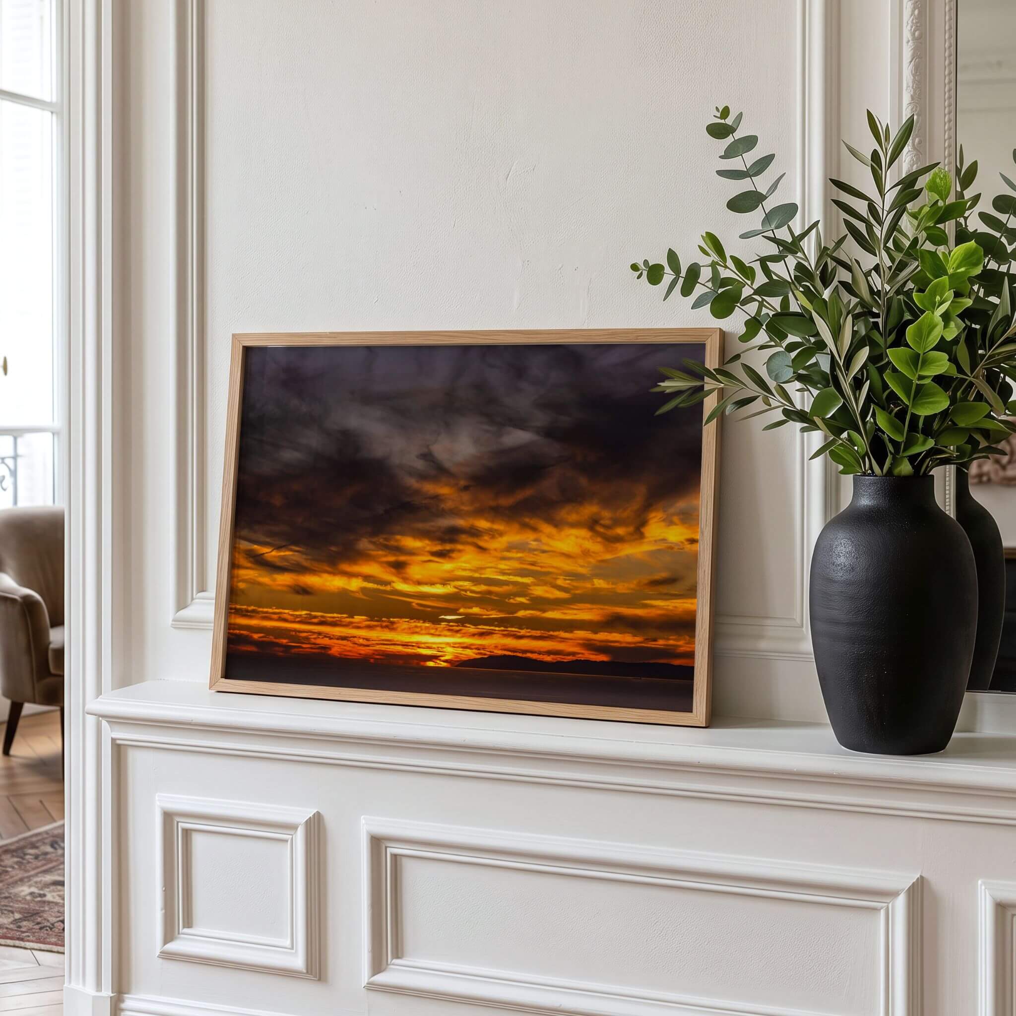 A framed wall art print featuring a dramatic sunset sky in orange, yellow, and dark gray tones, displayed on a white mantelpiece next to a black vase with green foliage.