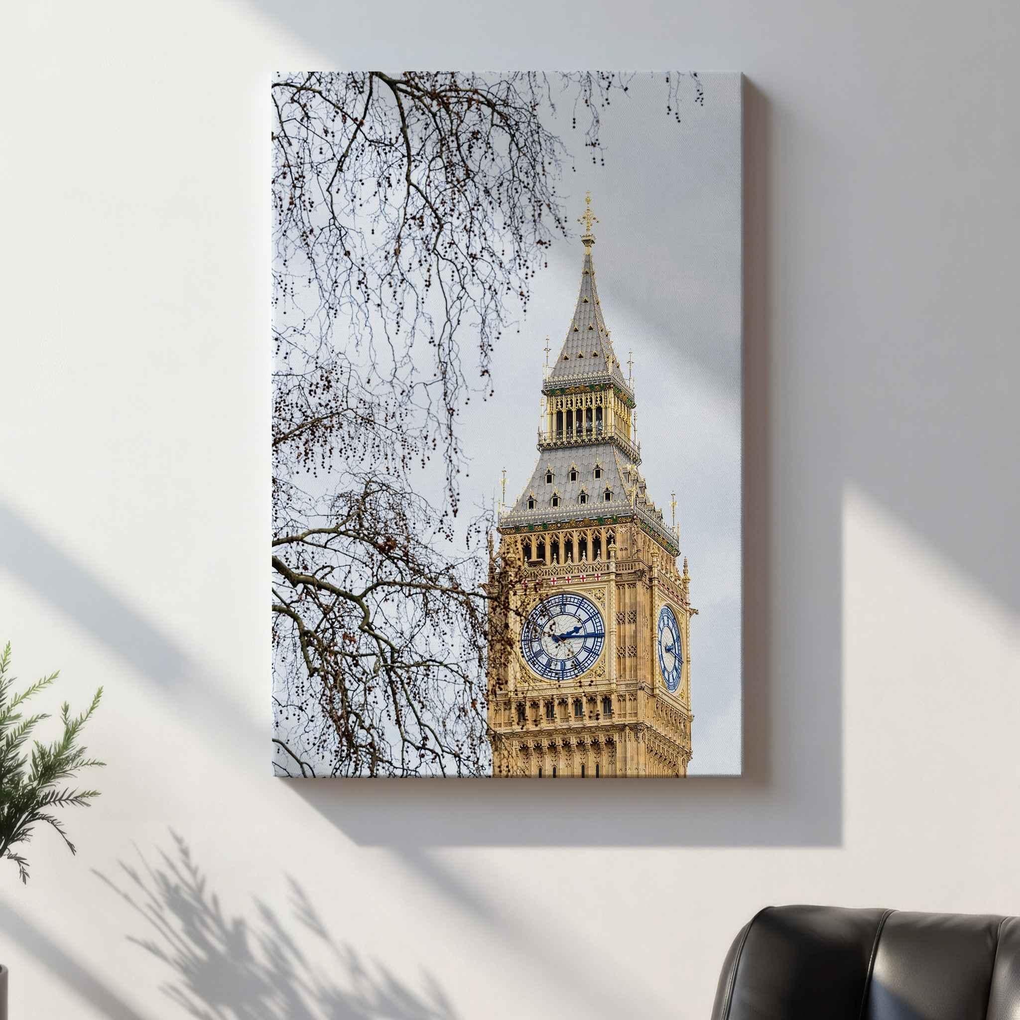A vertical canvas wall art print depicting Big Ben and the London Eye under a cloudy sky, with bare tree branches in the foreground. The gallery-wrapped canvas is shown hanging on a light gray wall with soft shadows, next to a dark leather chair and a small green plant.