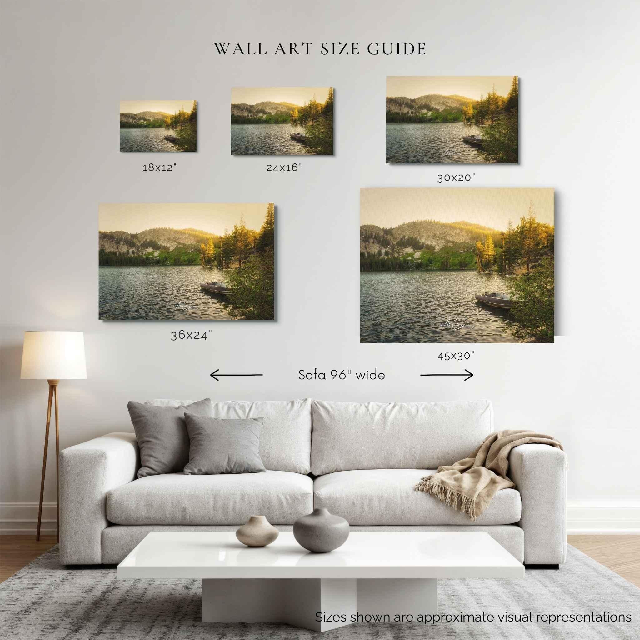 A wall art size guide displayed above a white sofa in a living room, showing five landscape canvas prints of golden hour alpine lake with dimensions labeled 18x12", 24x16", 30x20", 36x24", and 45x30". The canvases depict a lake scene at sunset with mountains, trees, and a small boat.