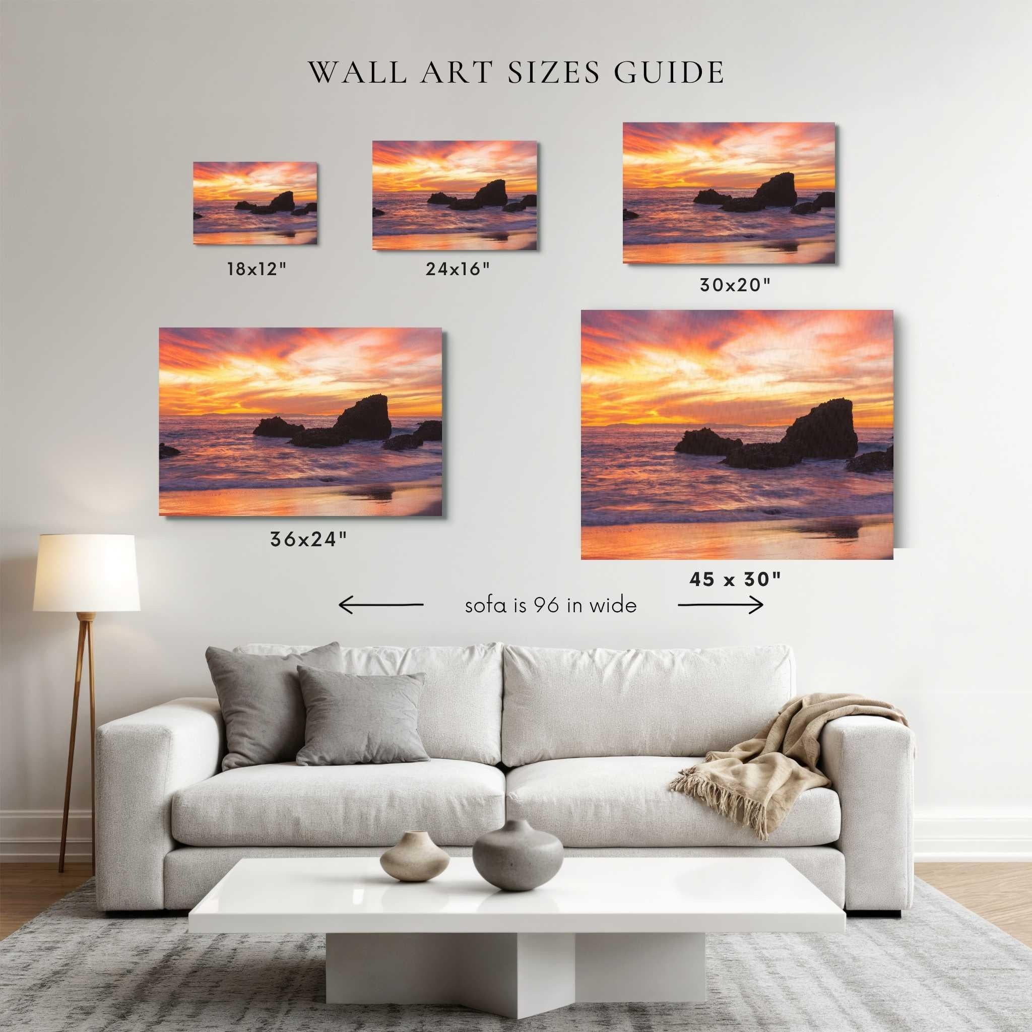 Five framed wall art prints showing ocean rock cliffs at sunset are displayed above a white sofa in a living room. Displaying sherbet sunset fine art gallery wrapped wall art in the sizes of each print: 18x12, 24x16, 30x20, 36x24, and 45x30.