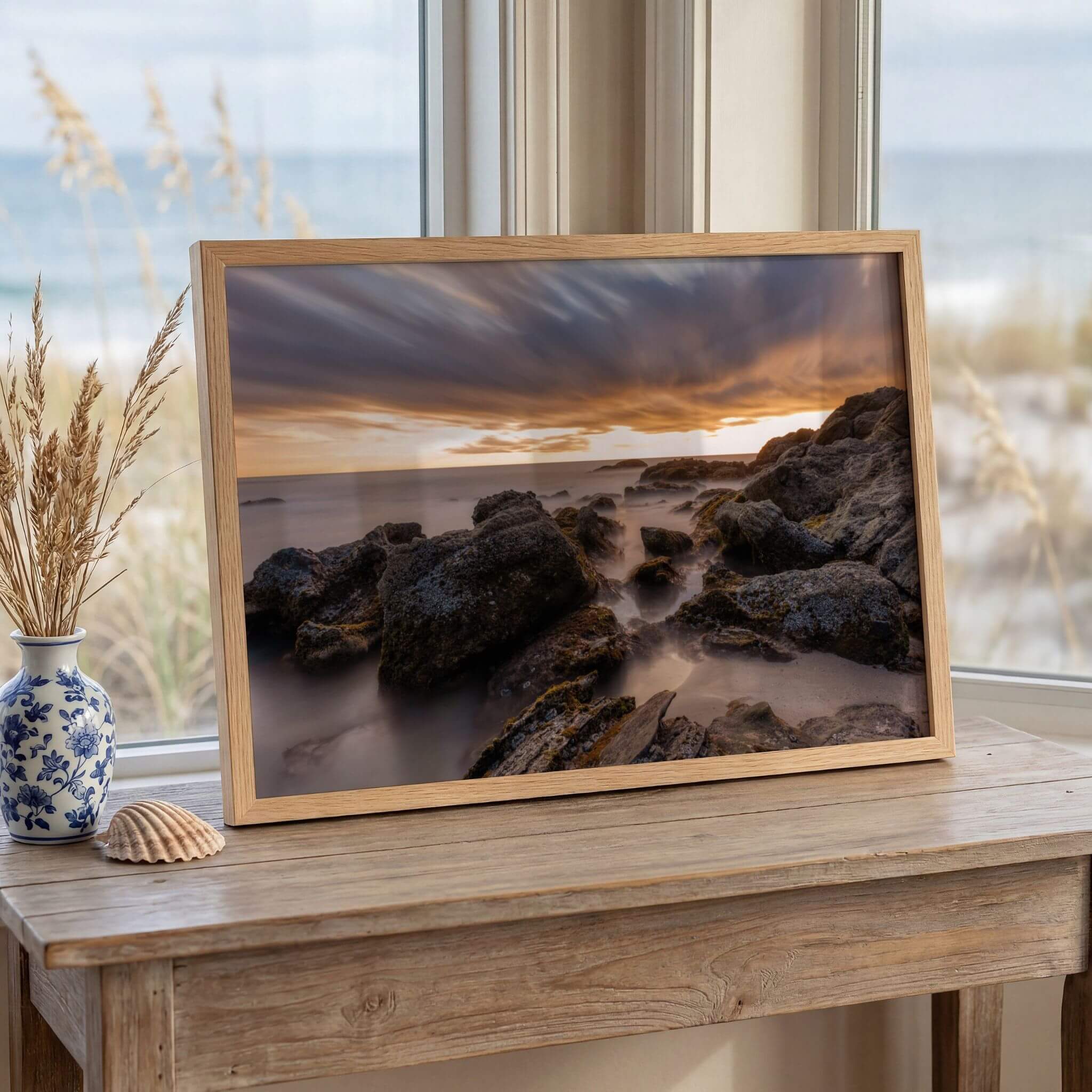 Laguna Beach coastal wall art displayed beside a beachside window, featuring dramatic sunset clouds, weathered shoreline rocks, and smooth long exposure ocean water.