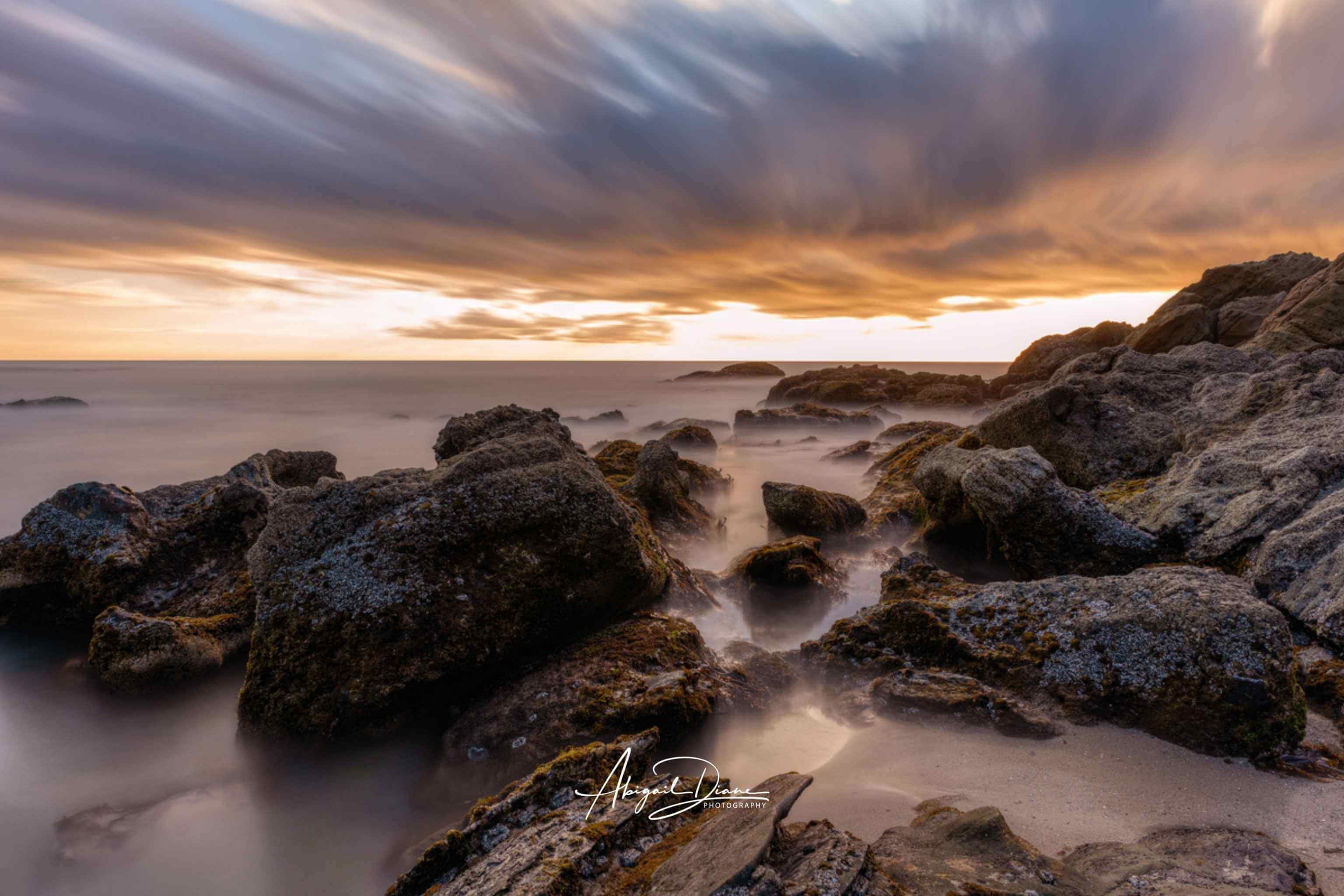 Long exposure coastal landscape photograph of Laguna Beach, California, featuring dramatic sunset clouds, rugged shoreline rocks, and silky ocean water.