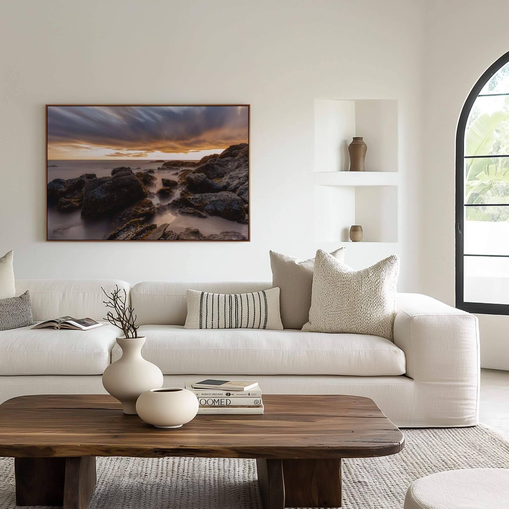 Long exposure Laguna Beach coastal wall art displayed above a modern white sectional sofa, featuring dramatic sunset clouds, rugged shoreline rocks, and silky Pacific Ocean water.
