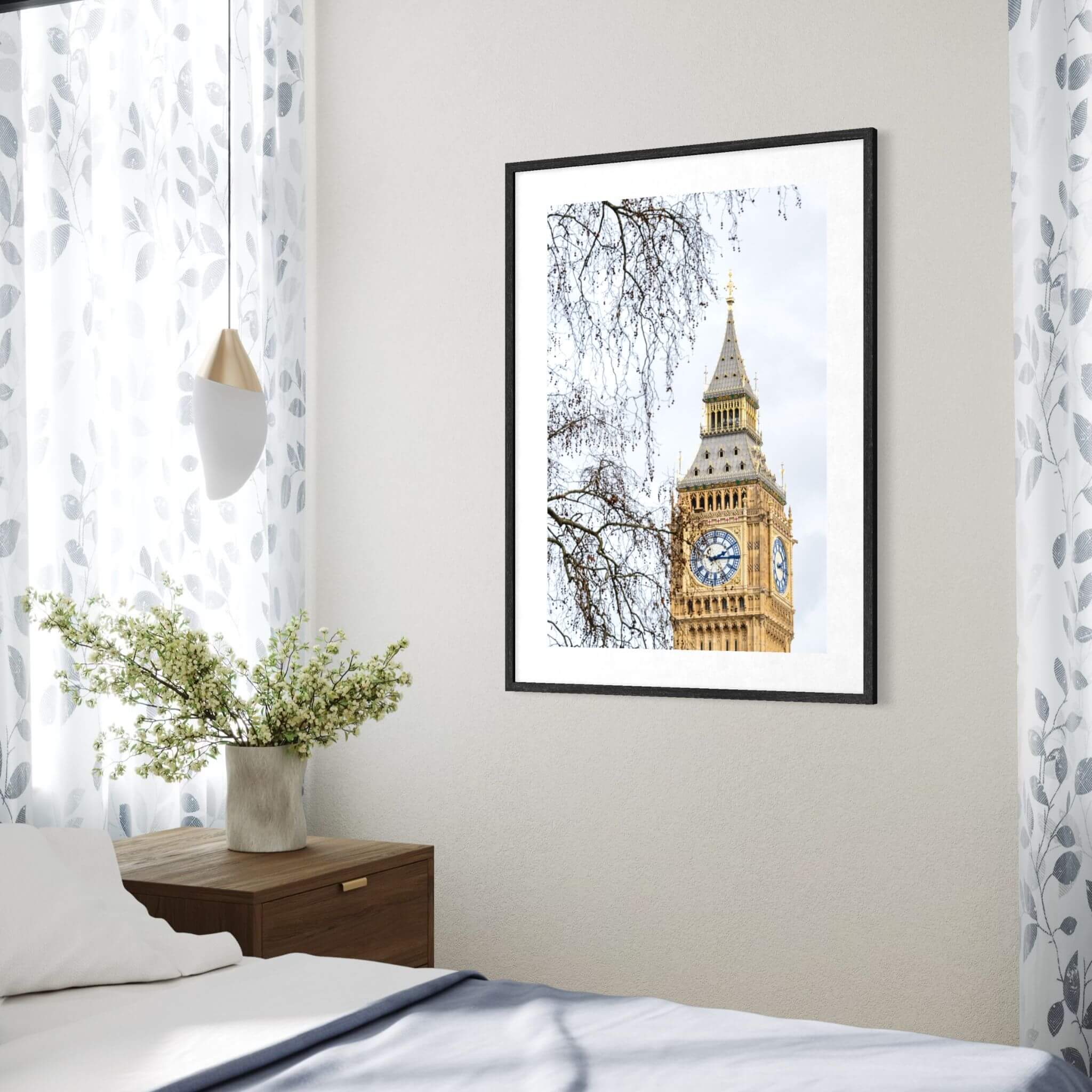 A framed vertical art print of Big Ben and the Elizabeth Tower in London, displayed on a light beige wall in a modern bedroom setting next to a wooden nightstand and patterned curtains.
