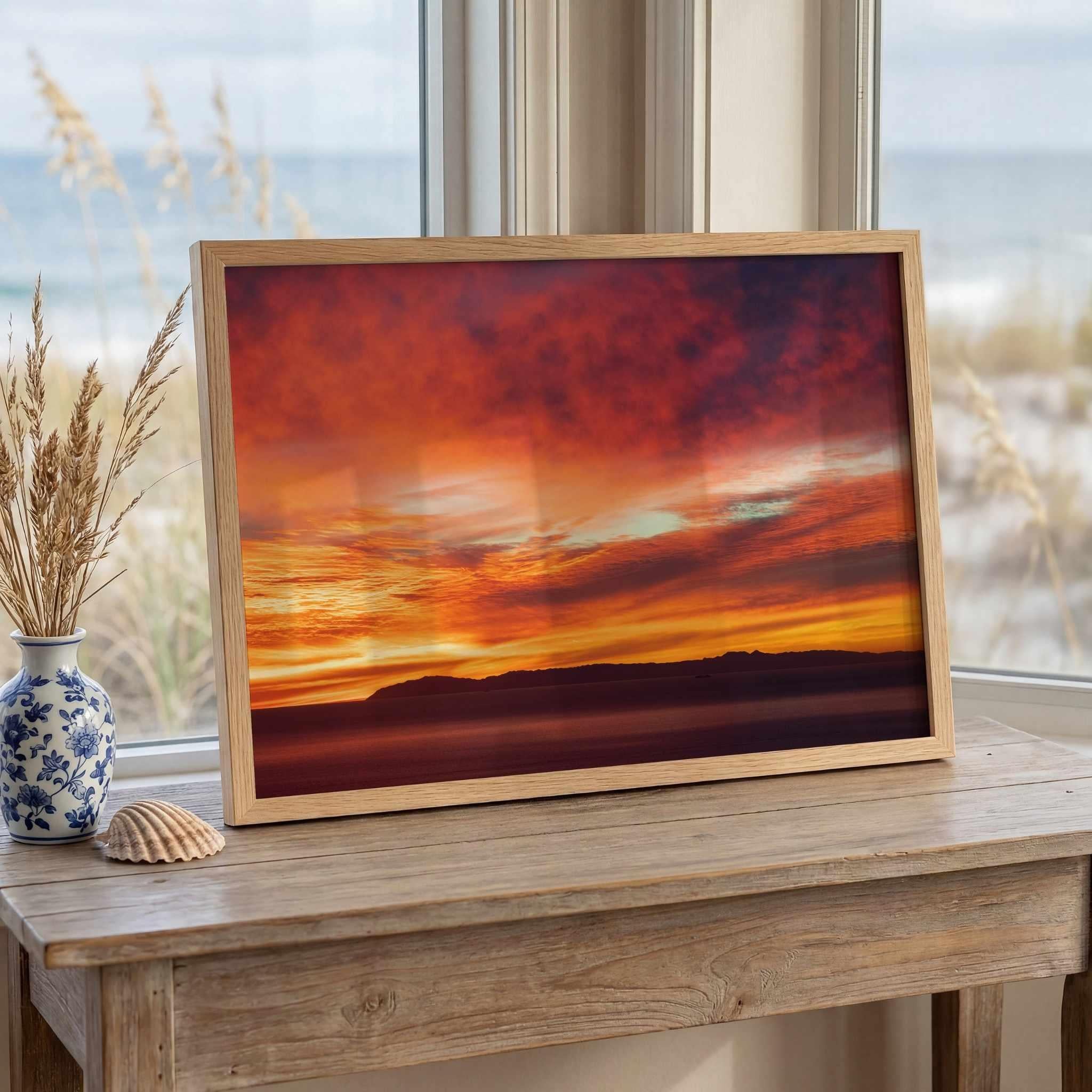Framed Catalina Beneath Crimson Skies fine art photography print leaning on a wooden console table beside a window with coastal décor, highlighting a vivid Catalina Island sunset over the Pacific Ocean.
