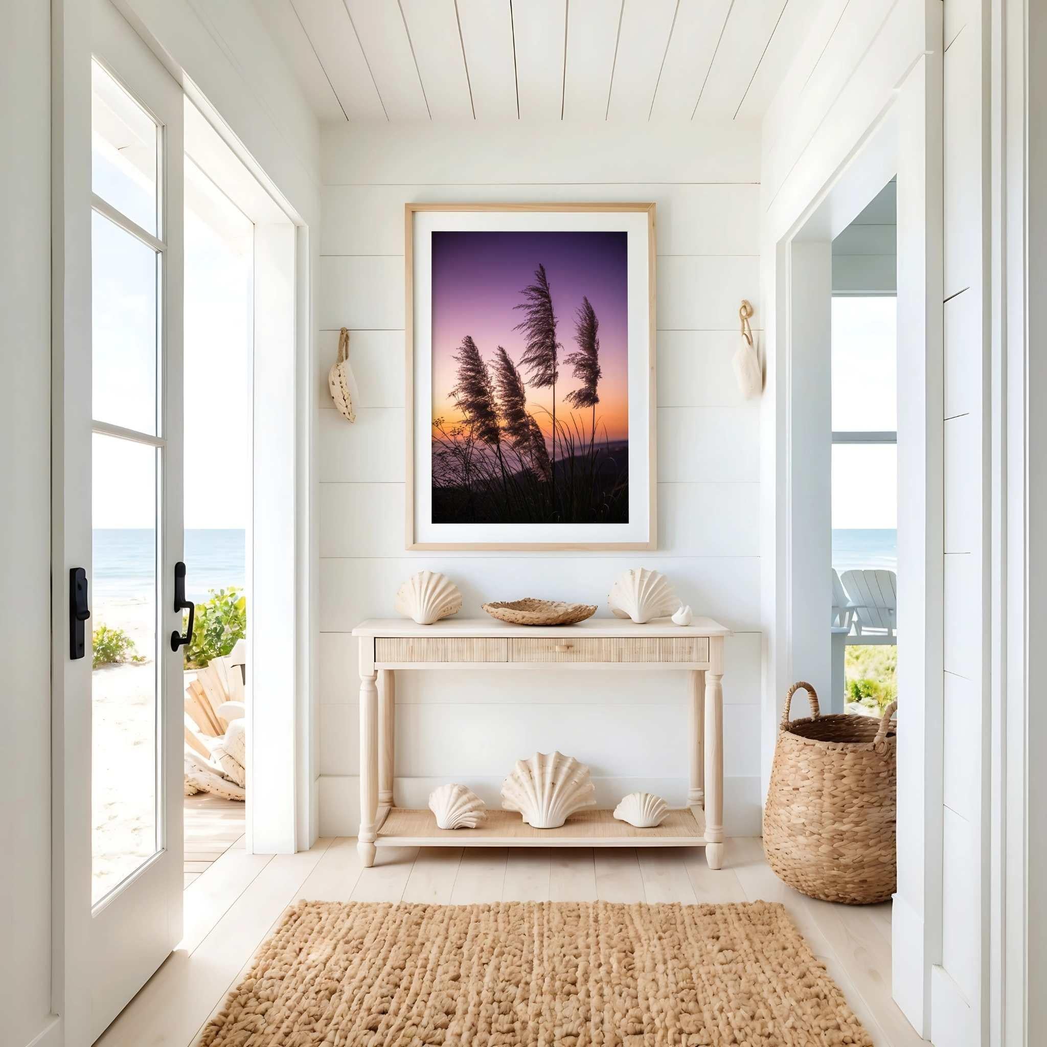 Fine art coastal pampas grass sunset photography by Abigail Diane Photography displayed in a light wood frame above a coastal console table. Soft neutral décor, seashell accents, woven textures, and ocean views create a bright beach house interior that complements the purple, pink, and golden hues of the sunset.