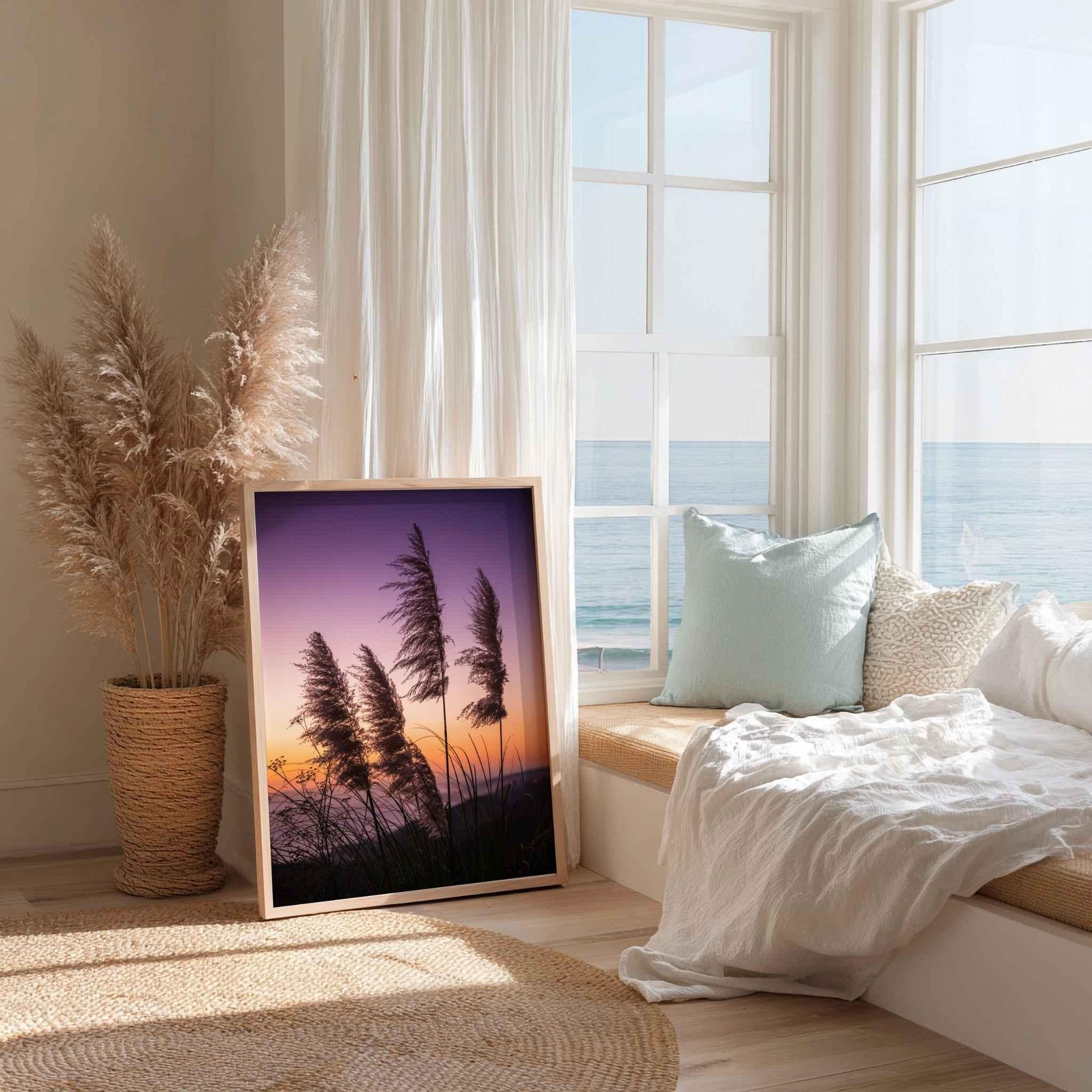 Fine art coastal pampas grass sunset photography by Abigail Diane Photography displayed in a light wood frame within a bright coastal-inspired reading nook. Soft white linens, natural textures, woven accents, and ocean views create a peaceful atmosphere that complements the purple, pink, and golden hues of the sunset sky.