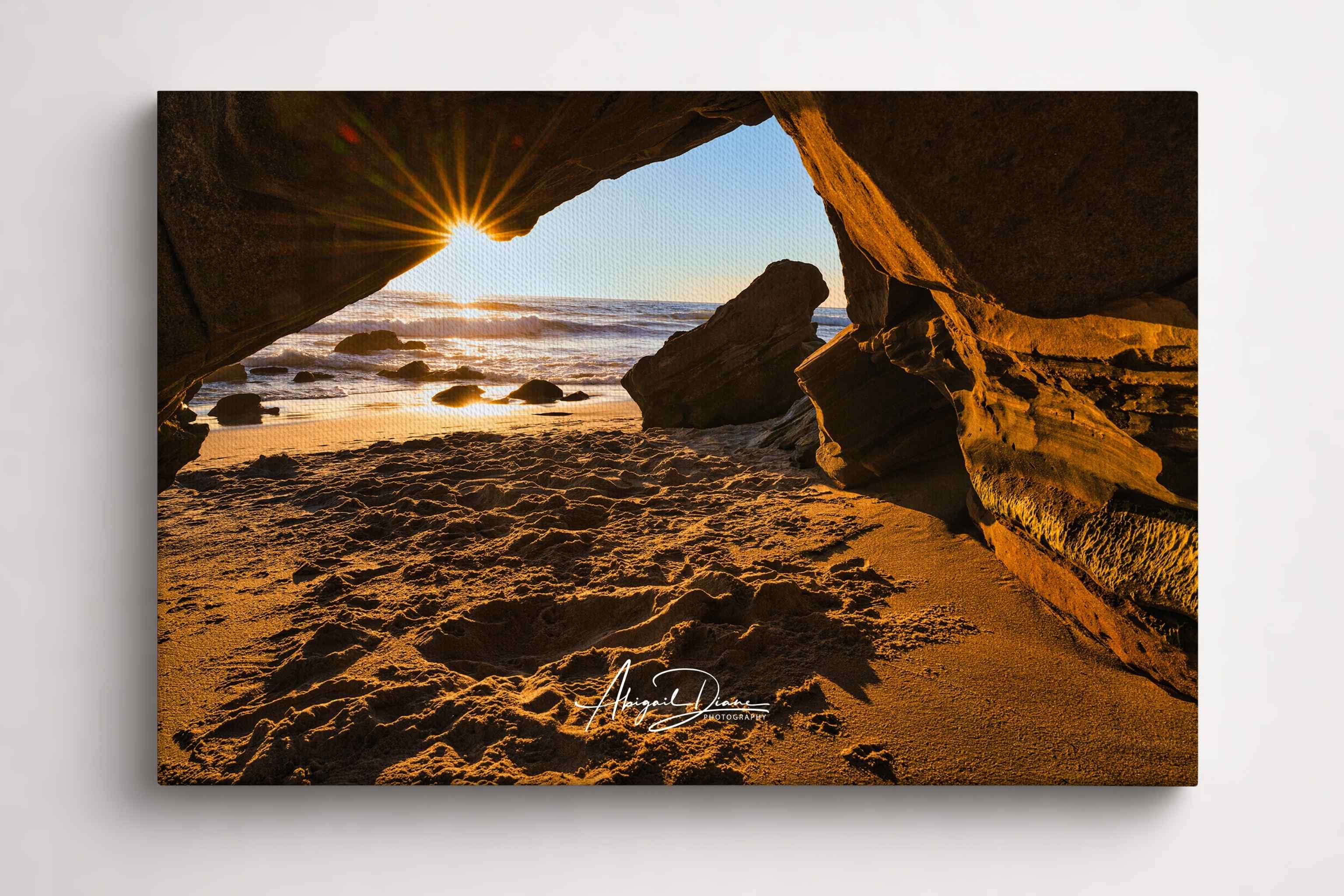Golden sunlight streams through a hidden sea cave at Laguna Beach, California, creating a radiant sunburst over the Pacific Ocean. Fine art canvas print by Abigail Diane Photography.