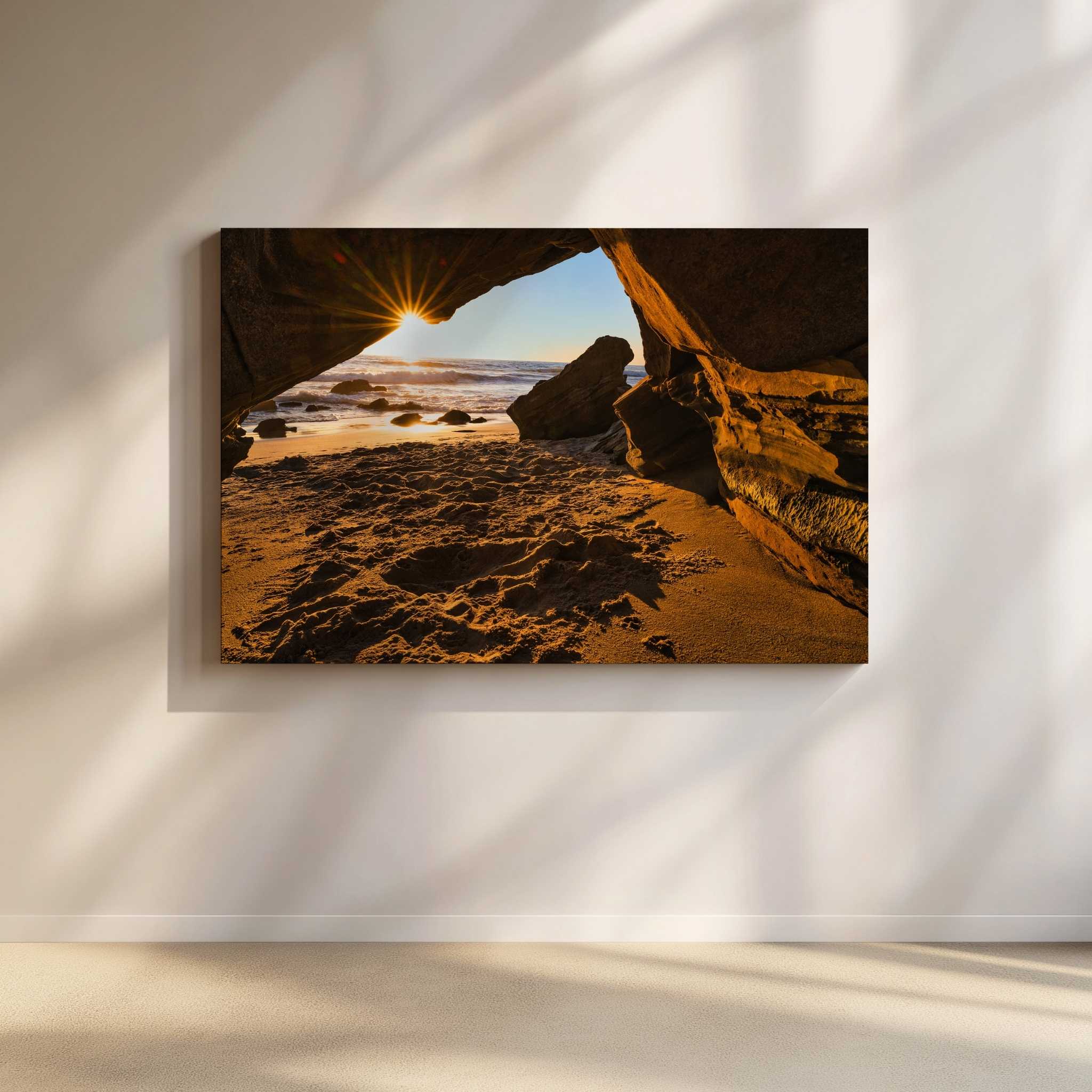 Golden Hour Sunburst fine art wall art featuring a radiant sunburst shining through a coastal sea cave at Laguna Beach, California. Warm golden light illuminates the sandstone rock formations, sandy shoreline, and Pacific Ocean, creating a dramatic sunset landscape photograph by Abigail Diane Photography.