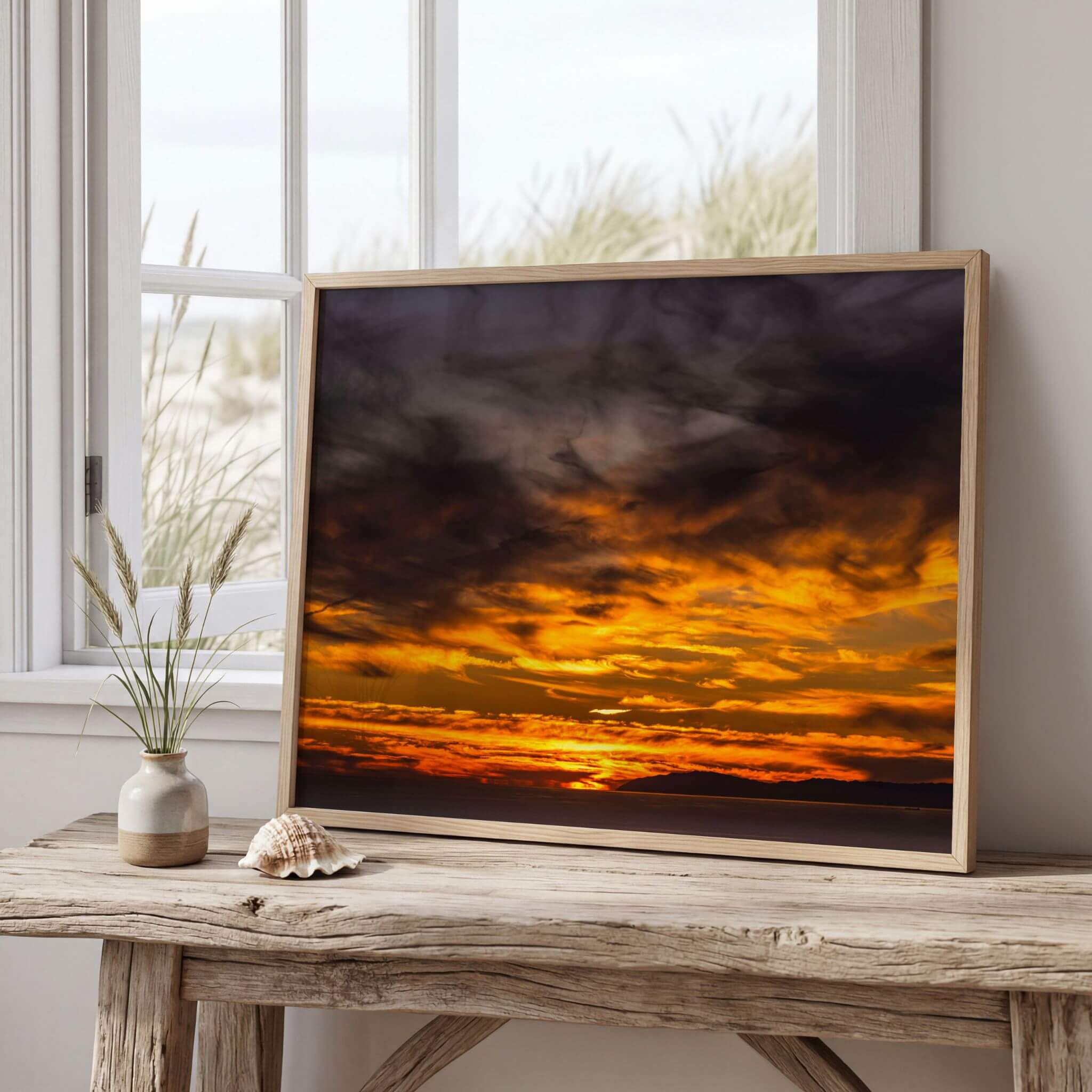 A framed golden sunset cloudscape art print depicting a dramatic orange and yellow sunset sky with dark clouds, leaning on a rustic wooden table next to a small vase with grass and a seashell.