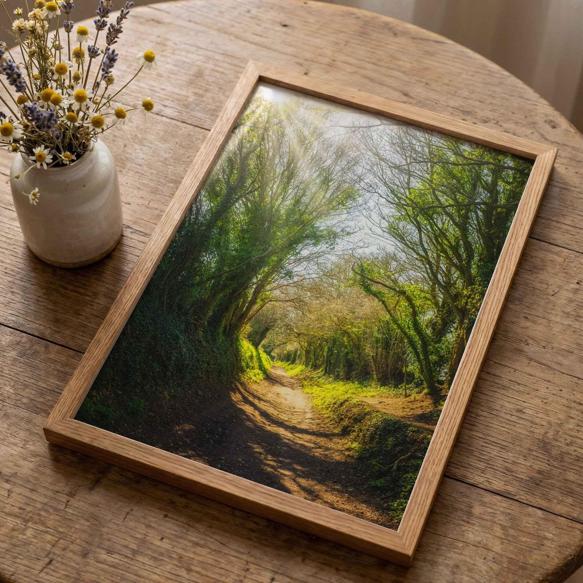 Framed Halnaker Tree Tunnel fine art print by Abigail Diane Photography resting on a rustic reclaimed wood table beside a ceramic vase of wildflowers. This historic tree-lined Roman road in West Sussex, England is showcased in a natural oak frame, highlighting the peaceful woodland landscape and timeless countryside beauty.