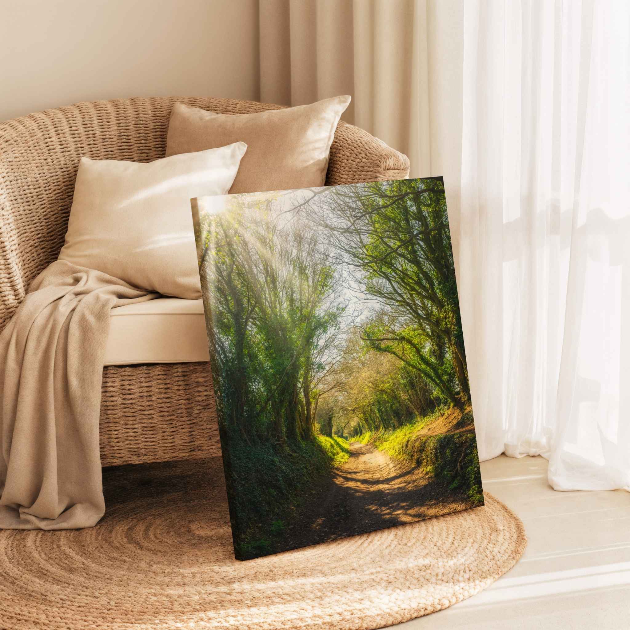 Wrapped canvas print of Halnaker Tree Tunnel by Abigail Diane Photography resting beside a woven rattan lounge chair with linen cushions, sheer curtains, and a natural jute rug. This peaceful woodland landscape from West Sussex, England brings warmth and tranquility to coastal-inspired home décor.