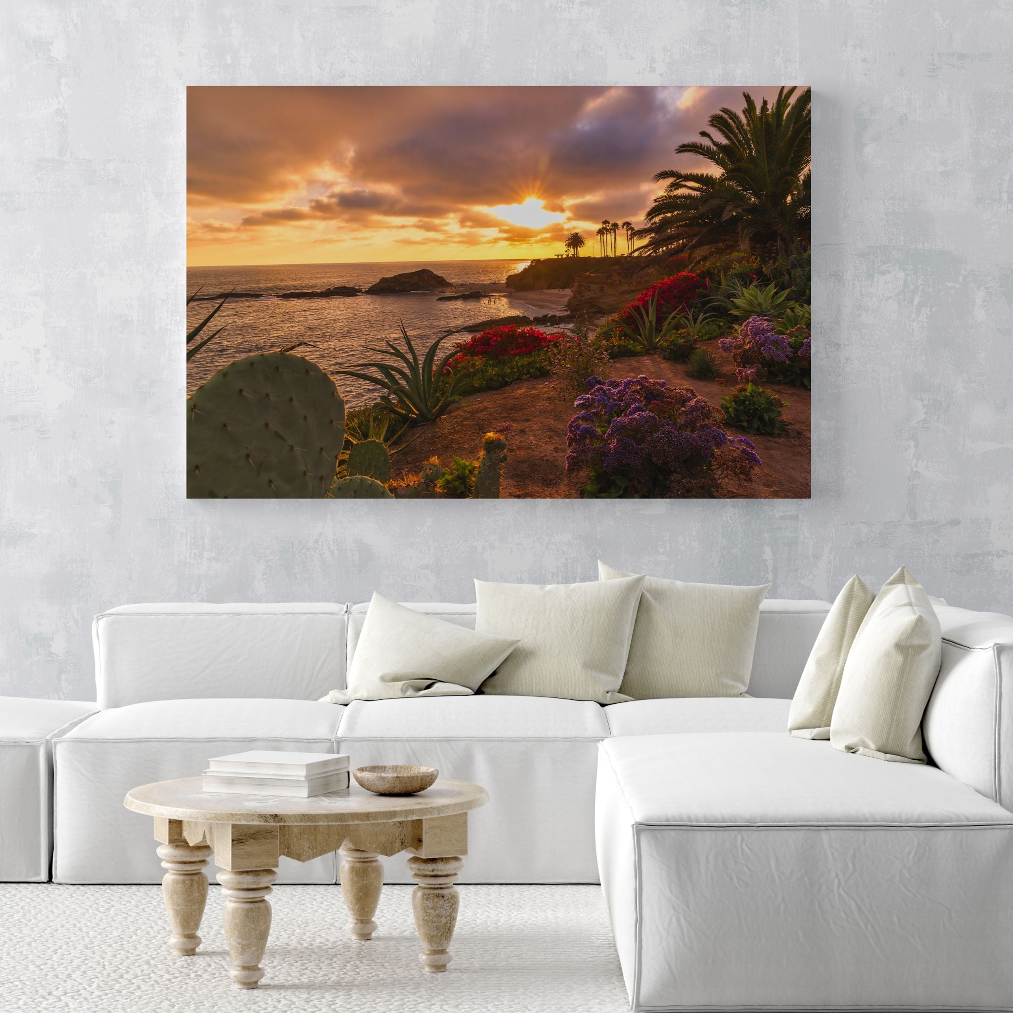 Gallery wrapped canvas featuring a Laguna Beach sunset with palm trees, coastal cliffs, and colorful flowers displayed above a white sectional sofa in a contemporary living room.