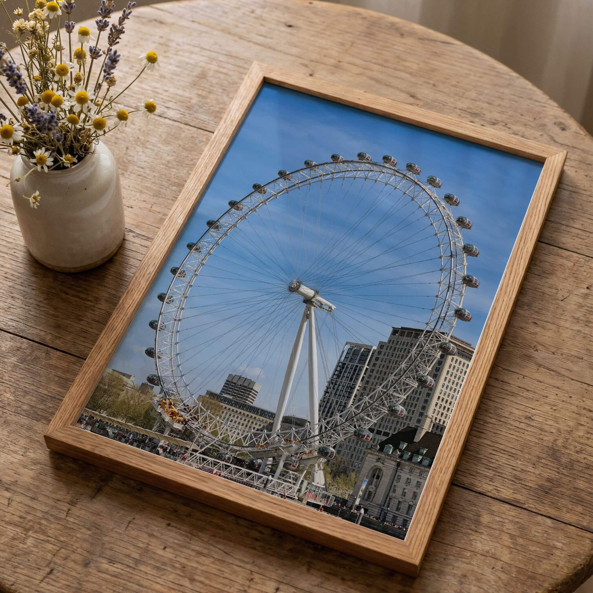 London Eye fine art print displayed in a natural wood frame on a rustic wooden table beside a vase of wildflowers.