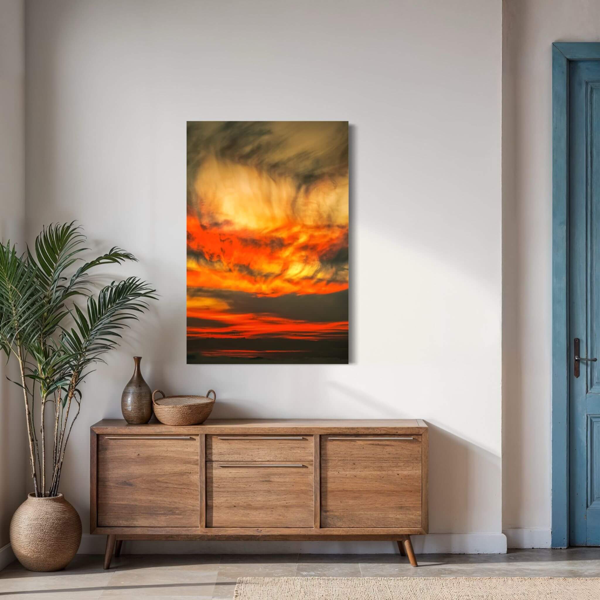Painted Lady High Definition metal print by Abigail Diane Photography, featuring dramatic golden yellow, fiery orange and deep charcoal sunset clouds, displayed above a natural wood console in a warm neutral interior with a blue accent door and indoor greenery.