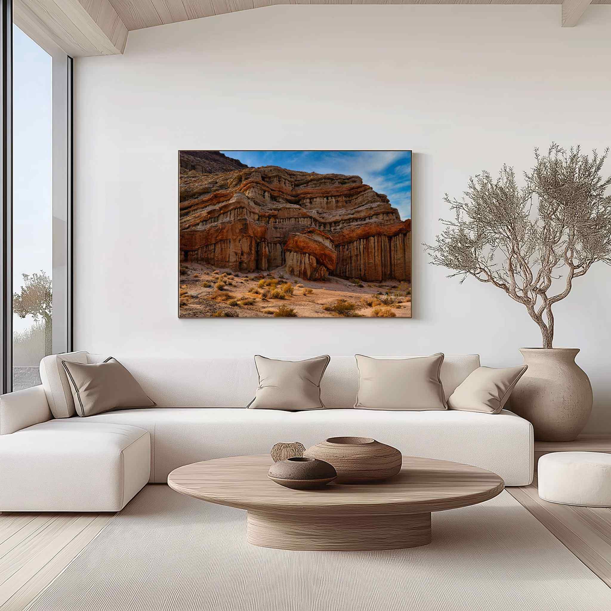 Large gallery-wrapped canvas of Red Rock Canyon State Park displayed above a cream sectional sofa in a modern organic living room. The artwork features California's dramatic layered sandstone cliffs, sculpted desert rock formations, warm desert earth tones, and a vivid blue sky, creating a striking statement piece for contemporary interiors. Artwork by Abigail Diane Photography.